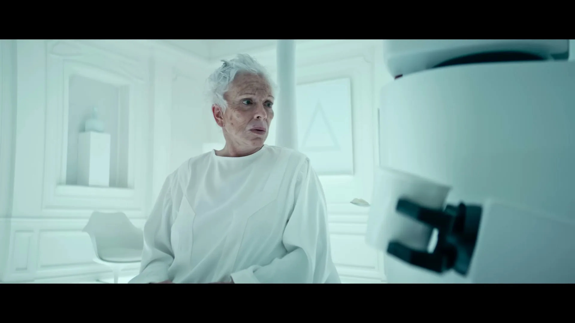 An elderly woman with white hair, wearing a white gown, looks with a worried expression at a robot in a sterile, all-white room.