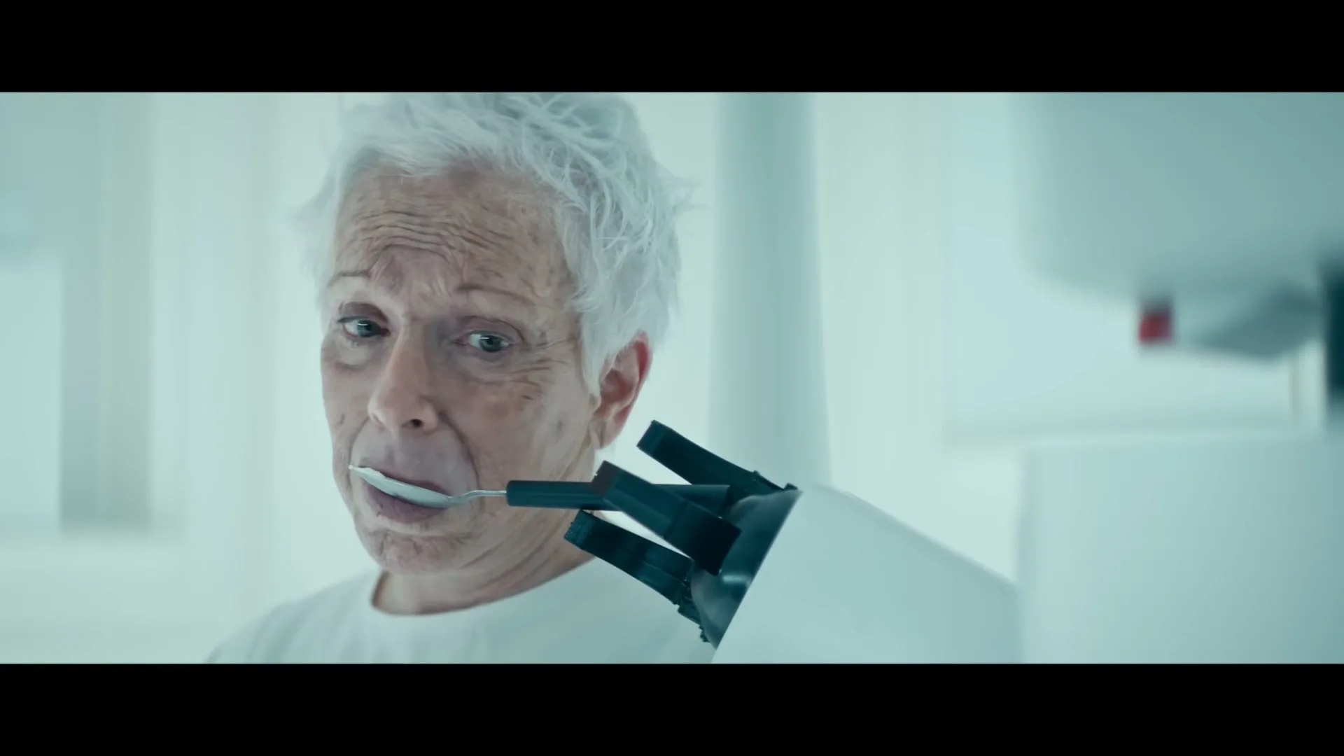 An elderly woman with white hair is fed a spoonful of ice cream by a robotic arm in a sterile, white room.