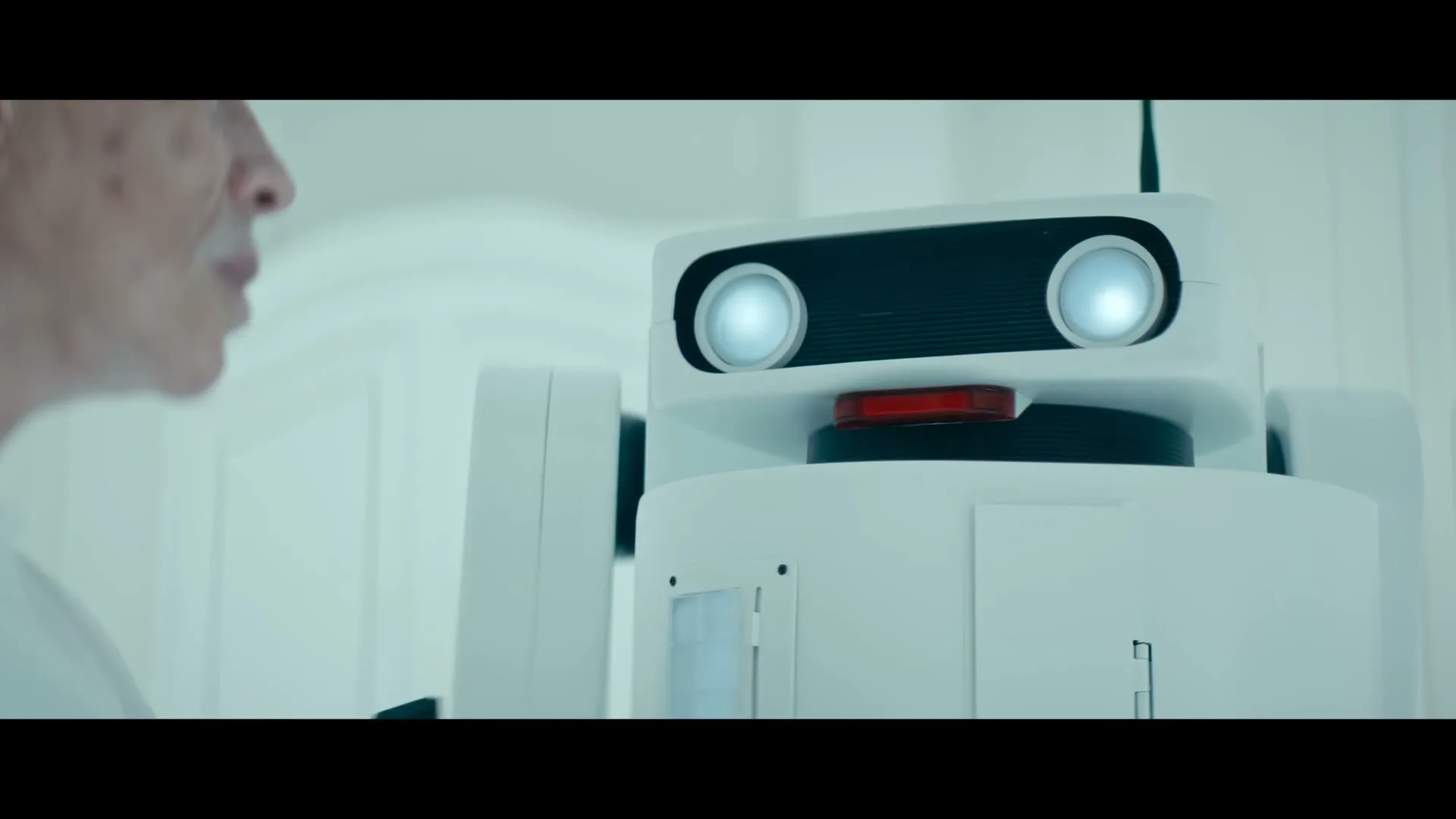 A pale woman with a robotic arm looks at a white robot with glowing eyes in a sterile, futuristic room.