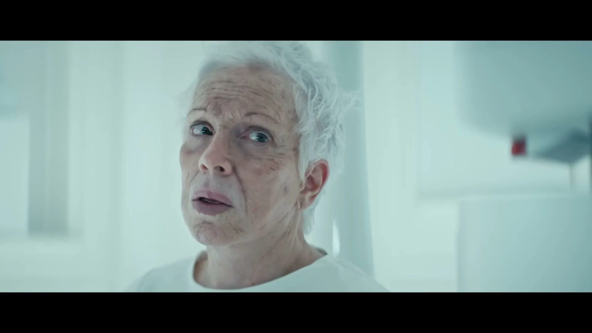 An older woman with white hair and a white shirt looks around with a confused expression in a sterile, white room.