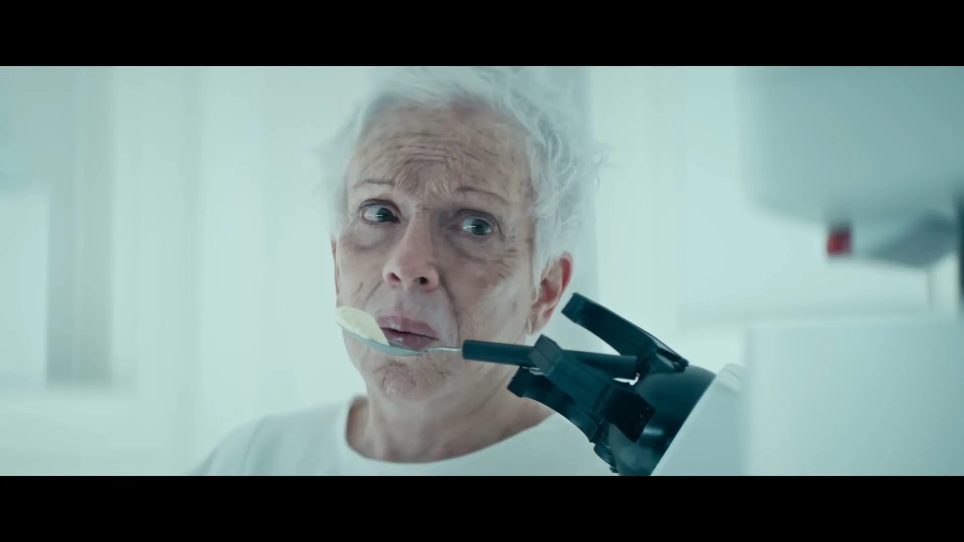 An elderly woman with white hair looks distressed as a robotic arm feeds her a spoonful of ice cream in a sterile, white room.