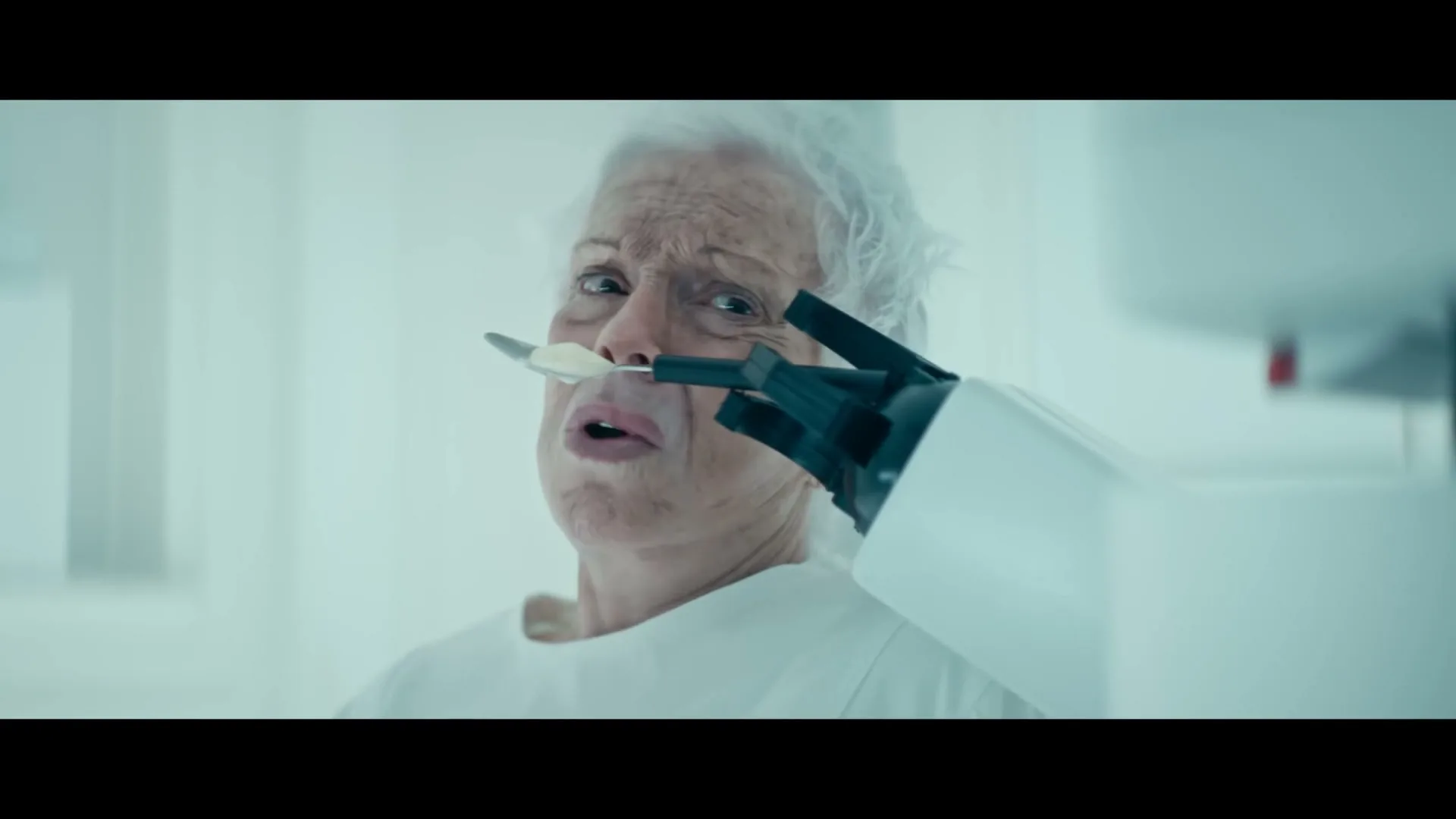 An elderly woman with white hair, wearing a white gown, is force-fed ice cream by a robotic arm in a sterile, white room.
