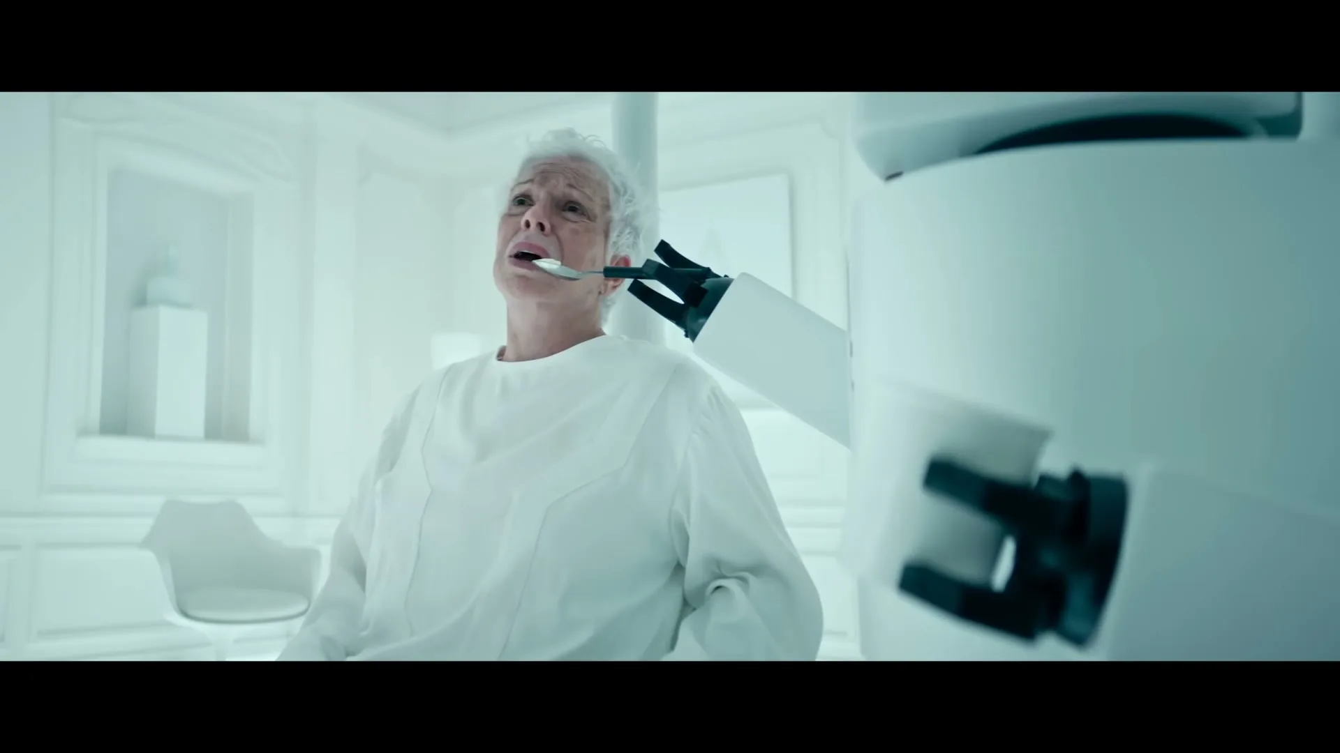 An older woman with white hair, wearing a white gown, is spoon-fed by a robotic arm in a sterile, all-white room, looking distressed.