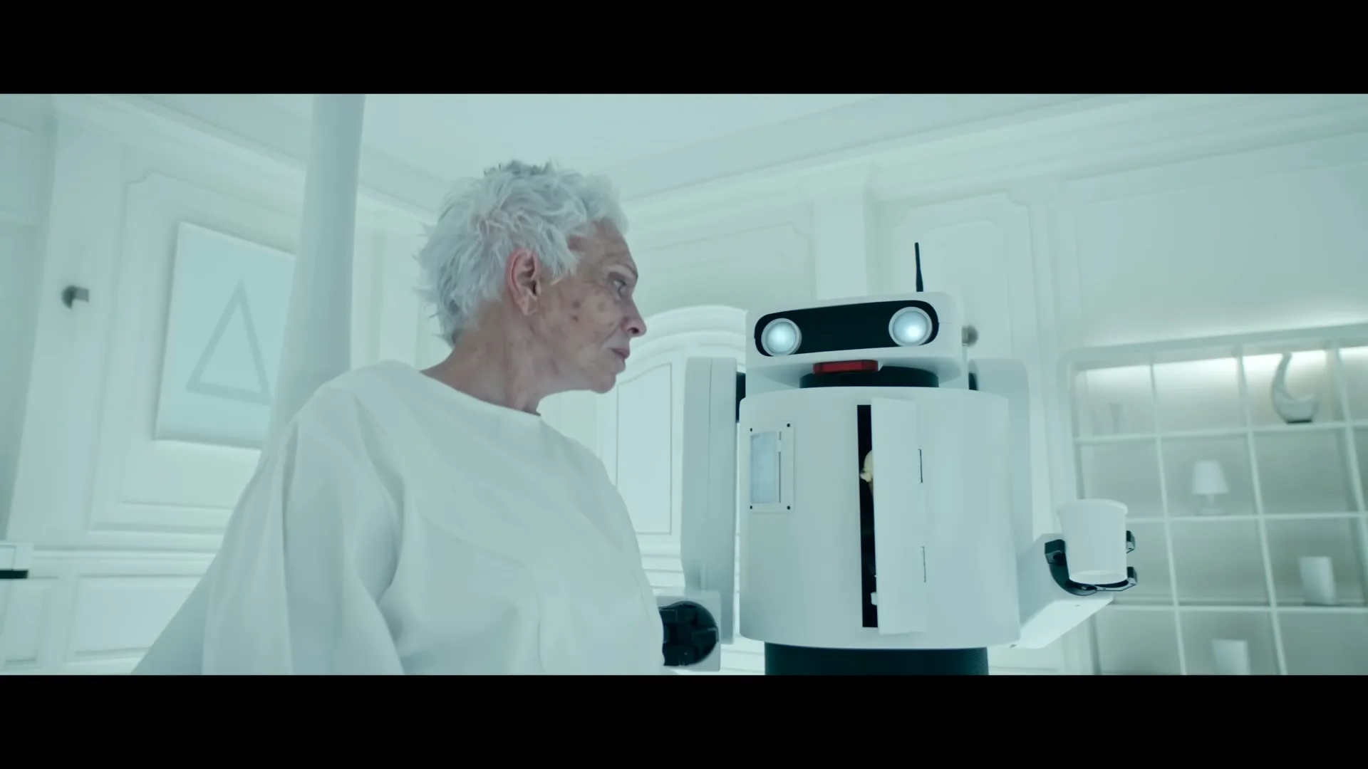 An elderly woman with white hair looks at a white robot that opens its chest to reveal an ice cream cone in a sterile, all-white room.
