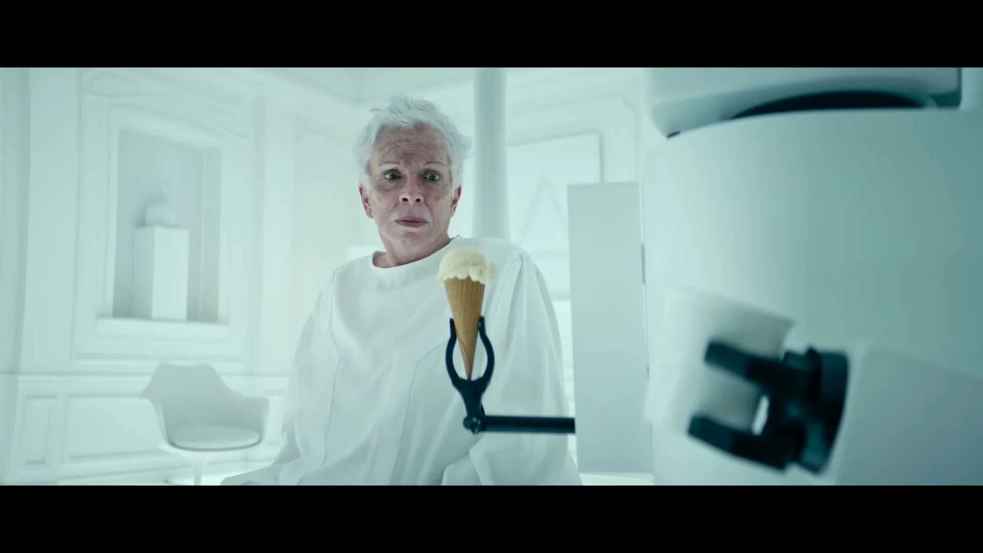 An older woman with white hair, wearing a white gown, looks directly at the camera with a worried expression while holding an ice cream cone in a sterile, all-white room.