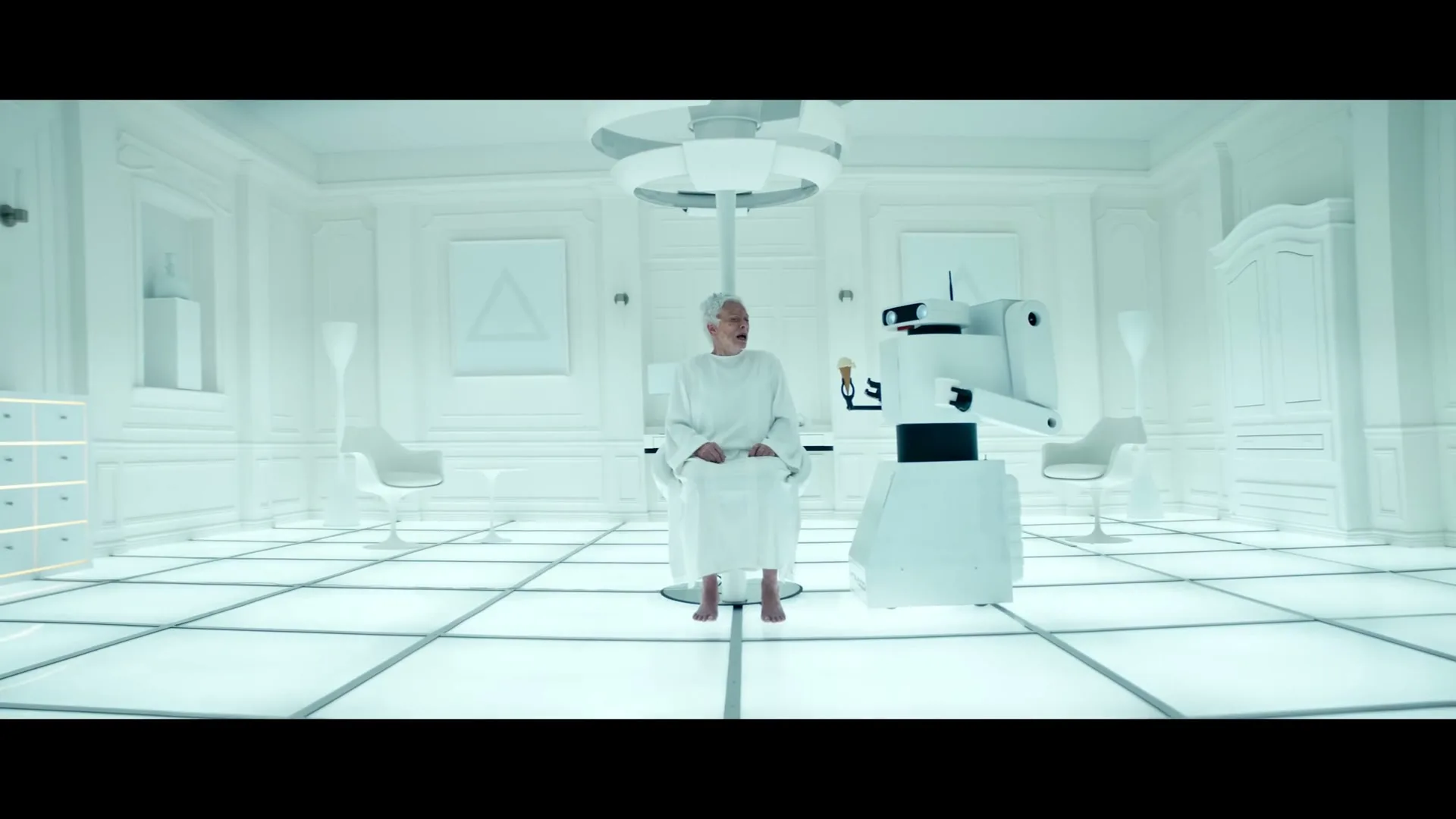 An elderly woman in a white gown sits in a futuristic, all-white room, looking at a robot holding an ice cream cone.