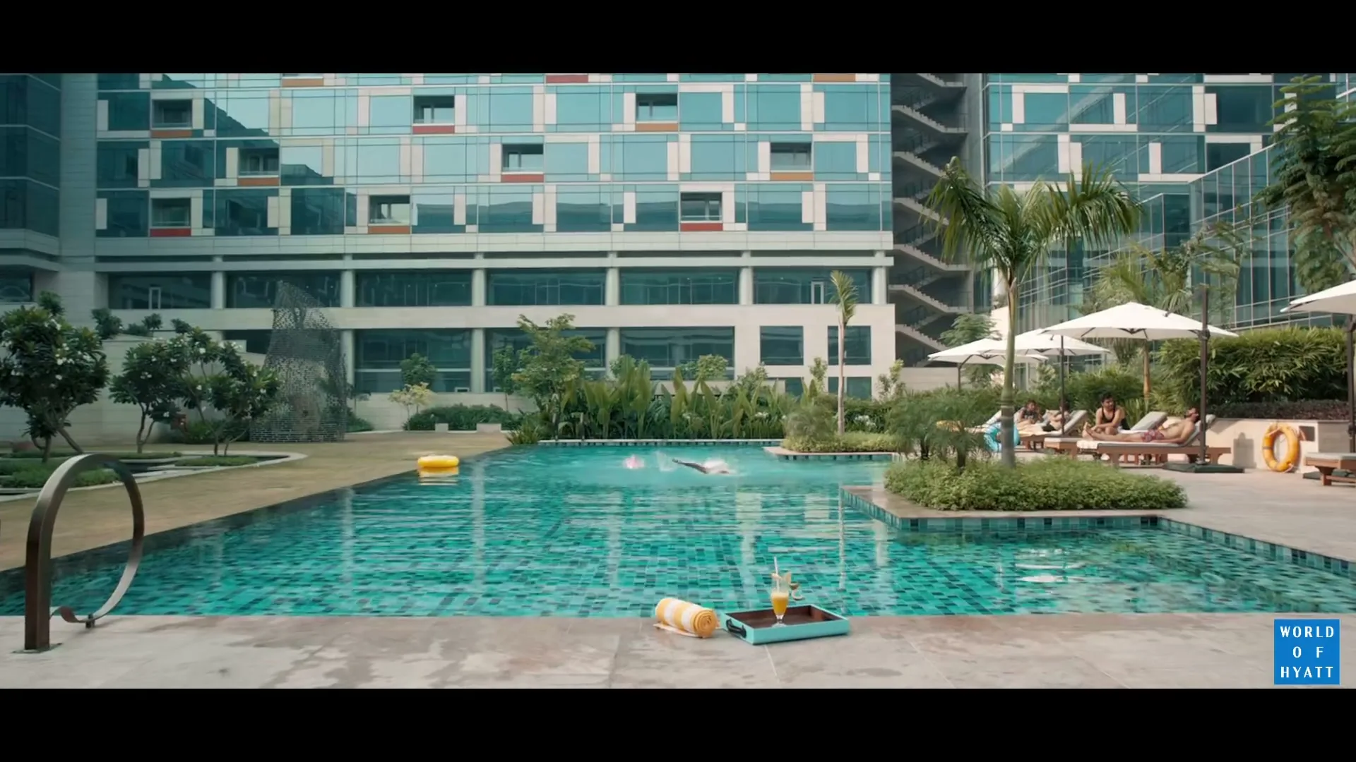 A wide shot of a modern hotel with a swimming pool, people swimming and relaxing by the pool on a sunny day.