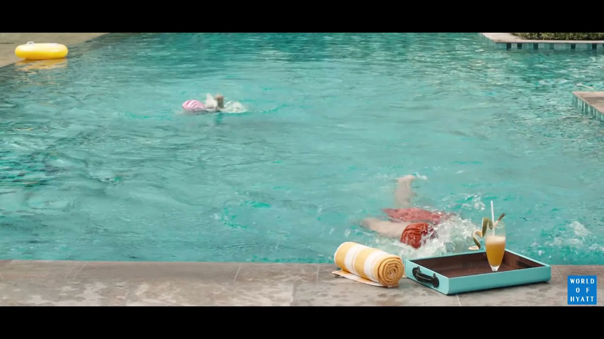 Two people swim in a hotel pool, one in a pink cap and the other in an orange cap, as a drink sits poolside.