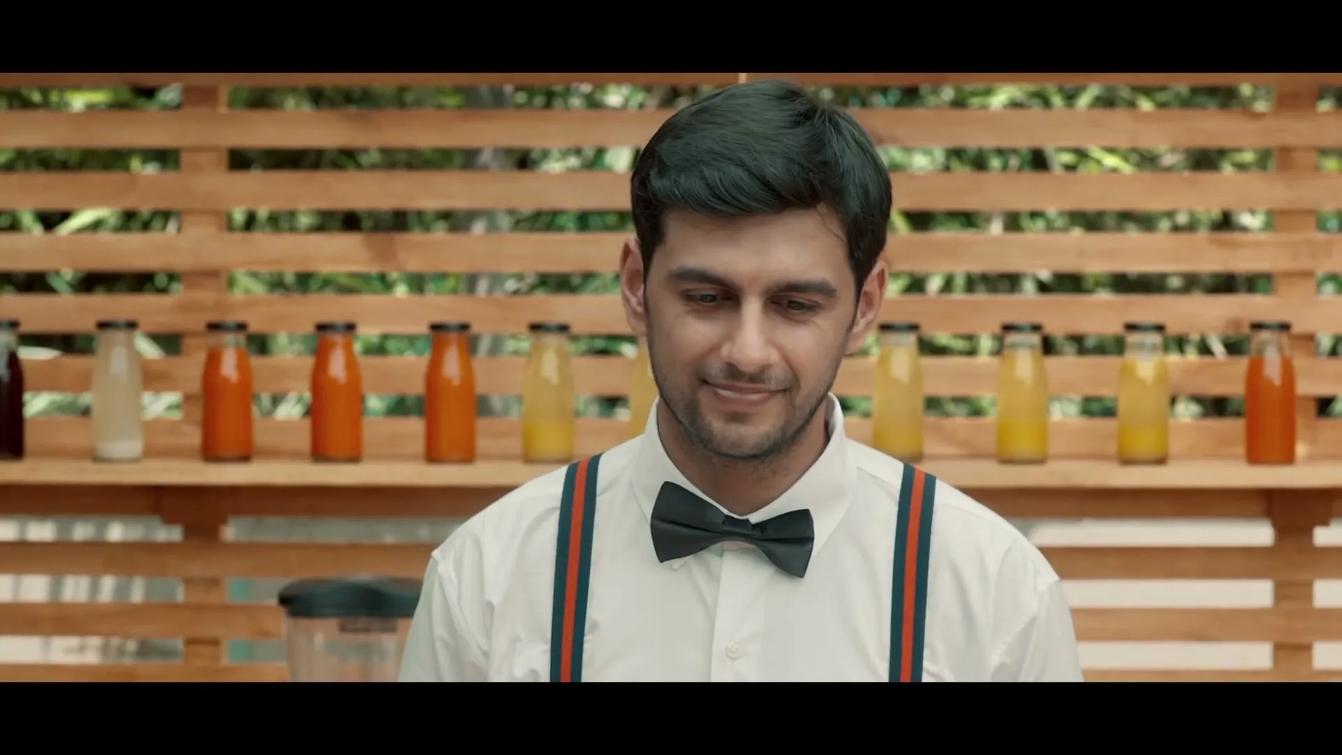 A young man with dark hair, wearing a white shirt and bow tie, smiles subtly while standing in front of a wooden slatted wall with bottles of juice.
