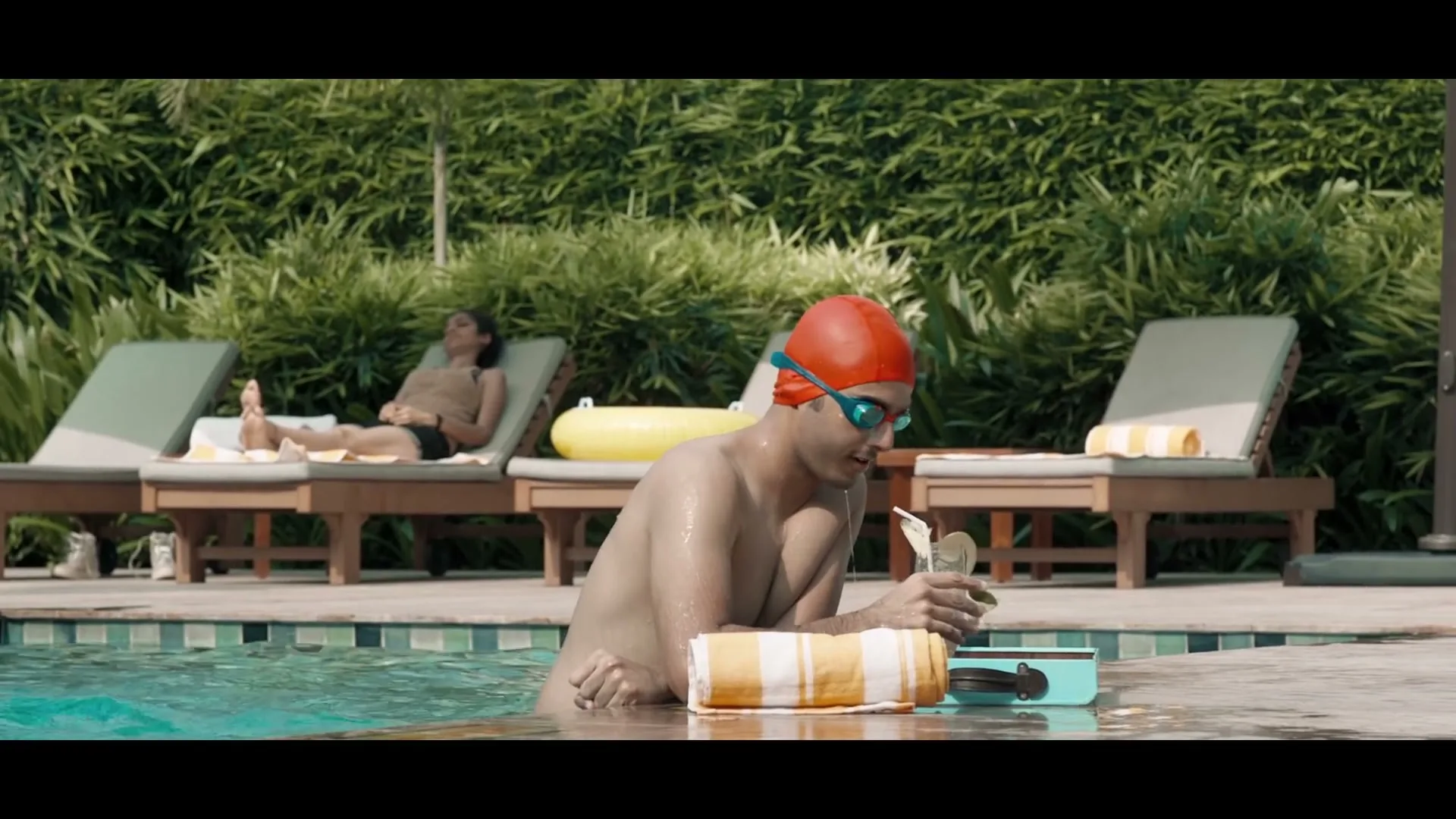 A man in a red swim cap and goggles is in a swimming pool, stirring a drink, while a woman sunbathes in the background.