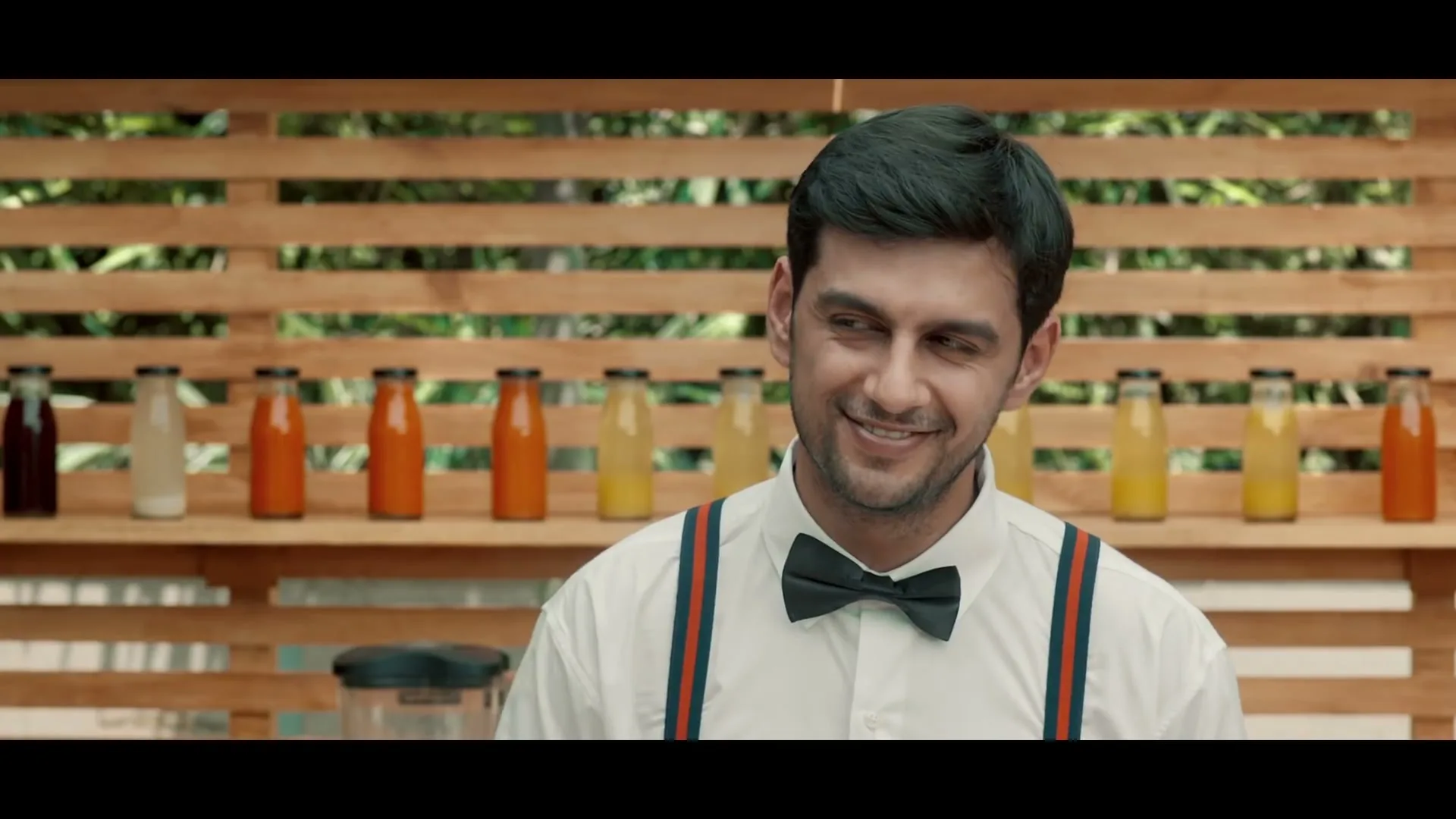 A young man with dark hair and a bow tie smiles while standing in front of a wooden wall with colorful bottled drinks on shelves.