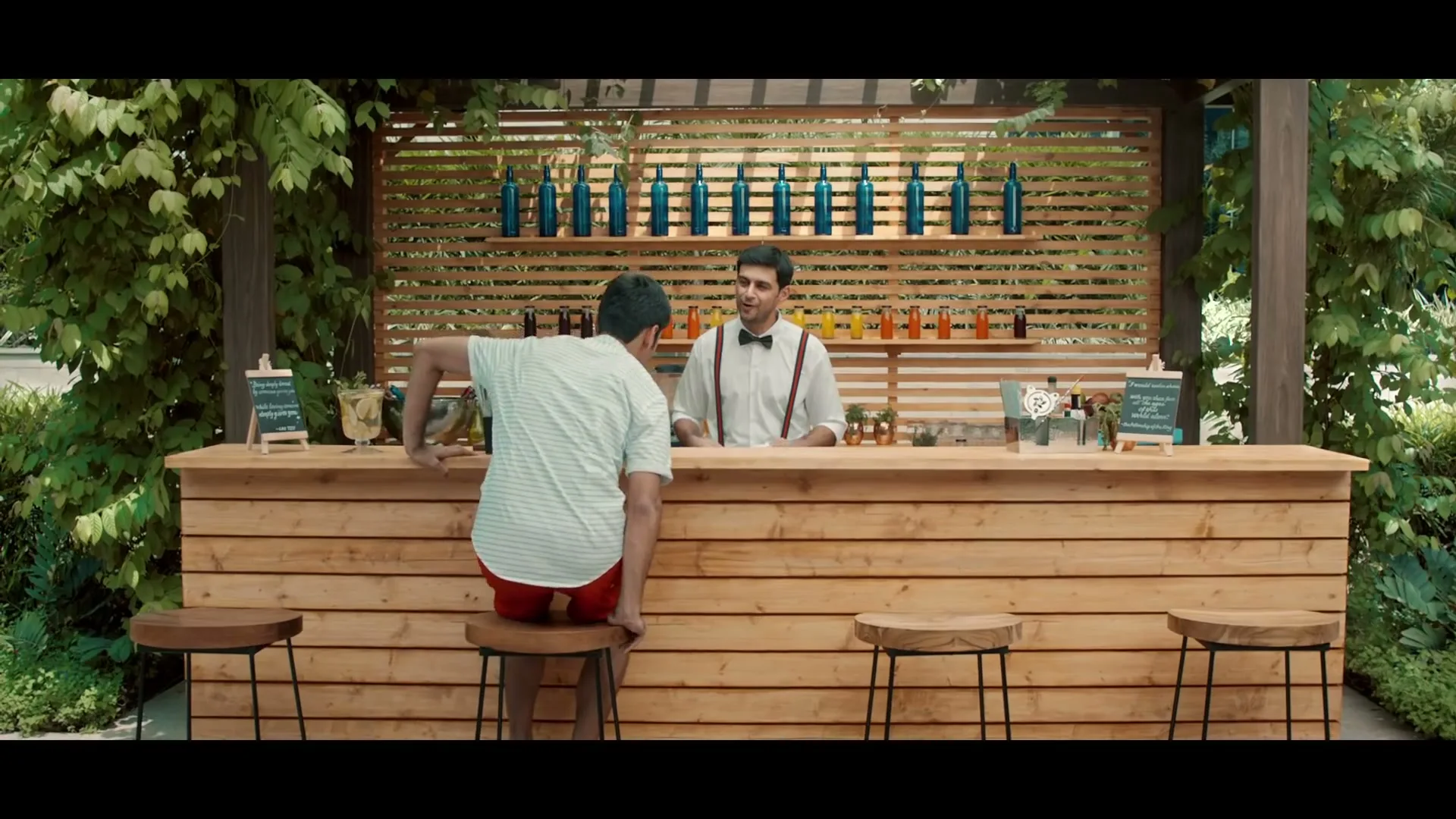A man in a white shirt and red shorts sits on a bar stool at an outdoor bar, talking to a bartender in a white shirt and bow tie.