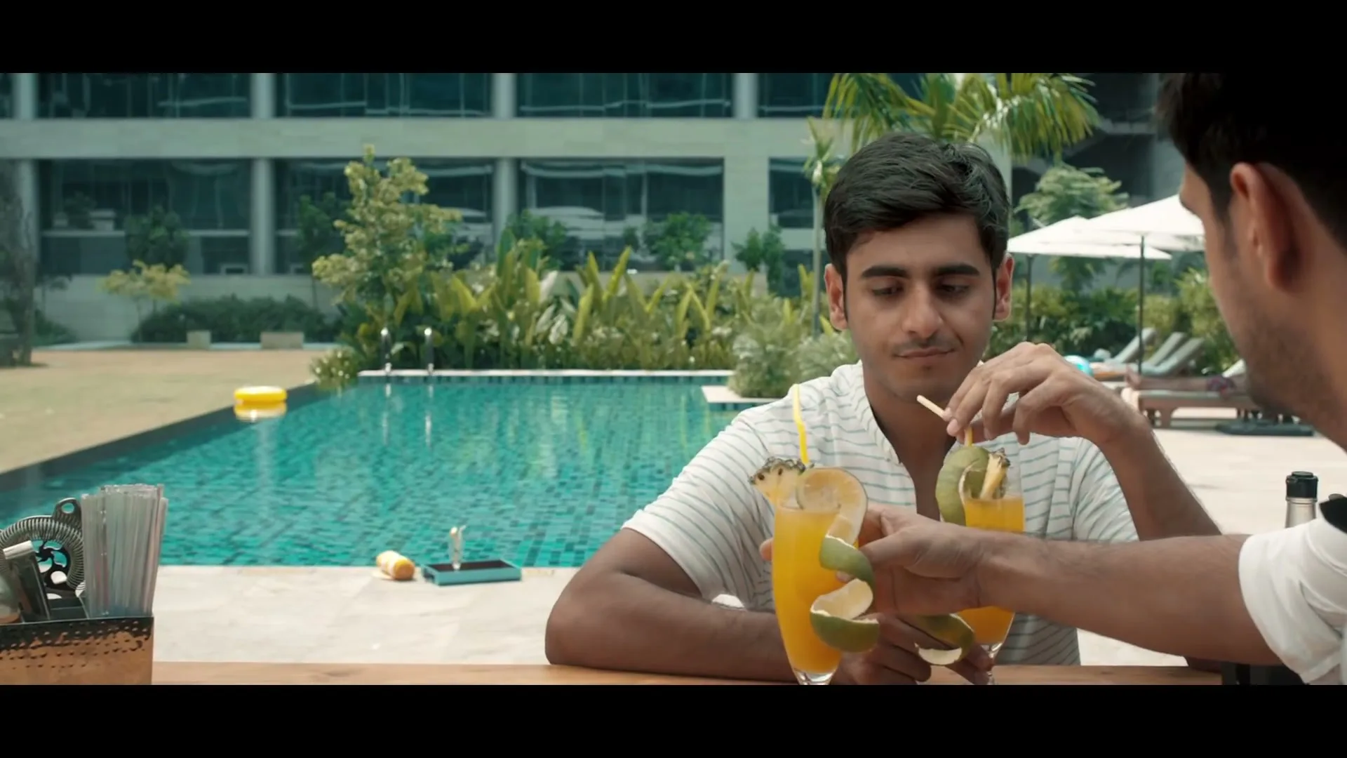 A young man in a striped shirt sips a cocktail at a poolside bar, then smiles as a woman in a swimsuit walks by.