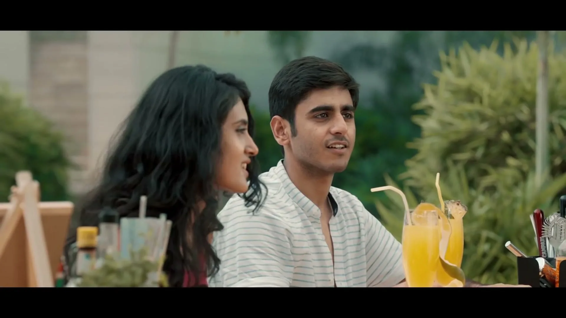 A young man and woman sit at an outdoor bar, talking and smiling, with tropical drinks in front of them.