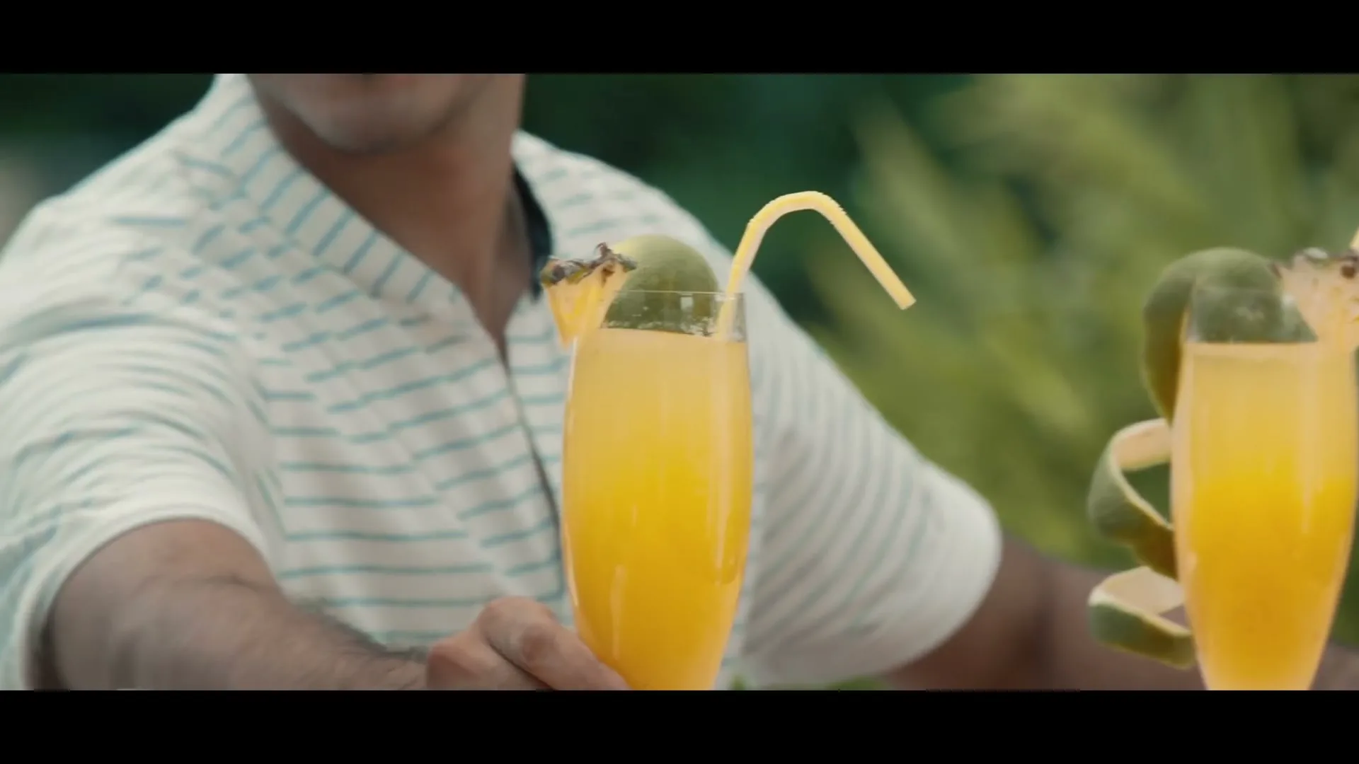 A man and a woman are sitting at a table outdoors, each holding a tall glass of orange juice with a straw and fruit garnish.