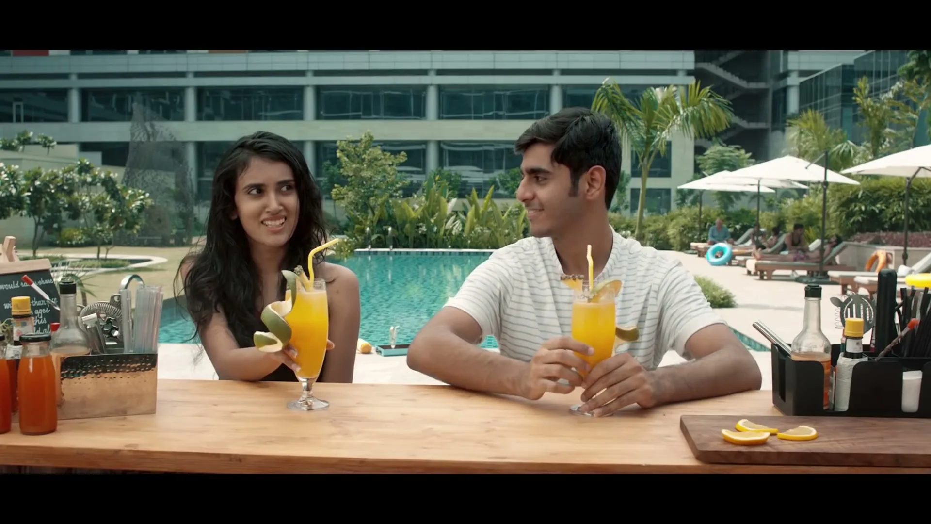 A young man and woman sit at a poolside bar, holding cocktails and smiling at each other, with a resort building in the background.