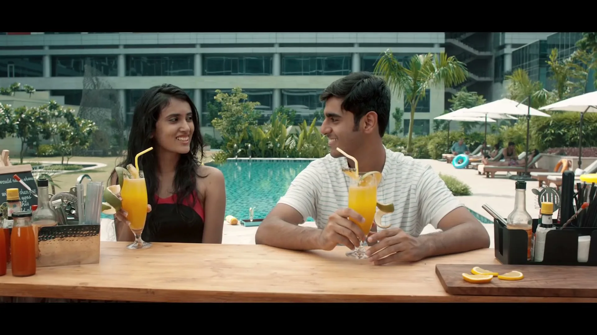 A young man and woman sit at a poolside bar, holding orange drinks with straws and garnishes, smiling at each other.