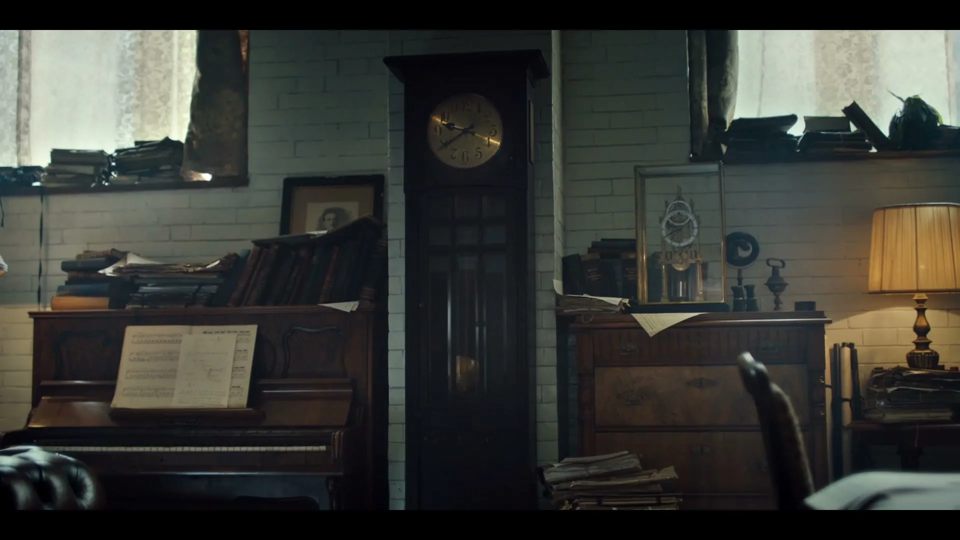 A wide shot of a cluttered, old-fashioned room with a grandfather clock in the center, a piano on the left, and a dresser on the right, all bathed in natural light.