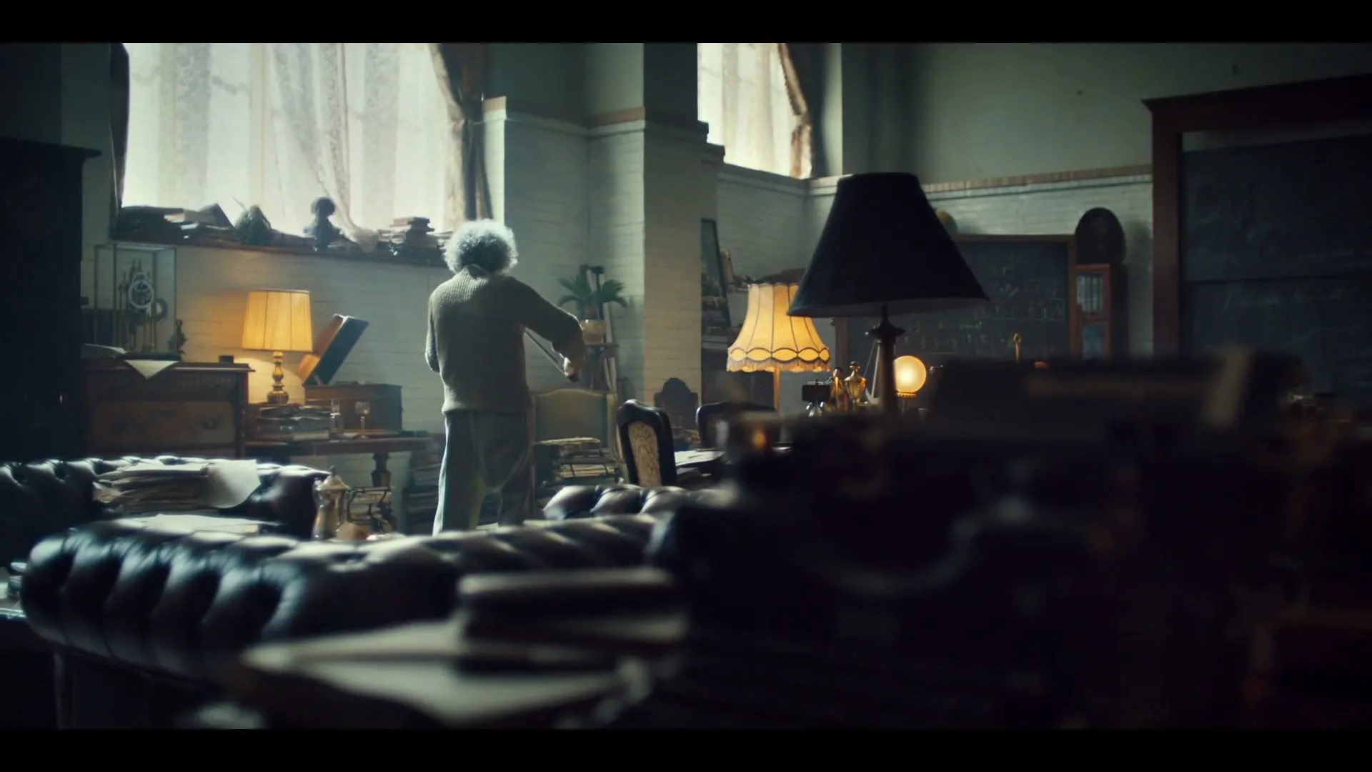 A man with wild white hair, dressed in a sweater and pants, plays a violin in a dimly lit, cluttered study with large windows.