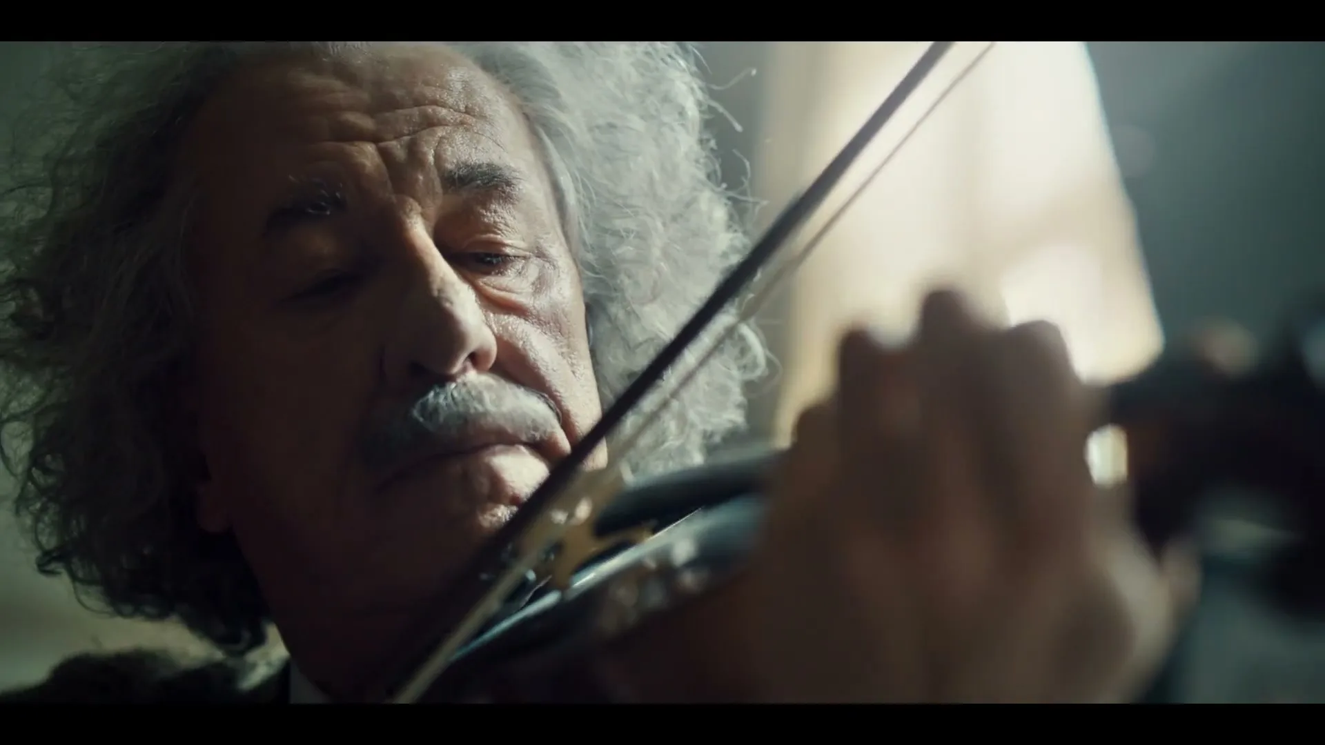 An older man with a mustache and wild grey hair, resembling Albert Einstein, plays the violin with a focused expression in an interior setting.