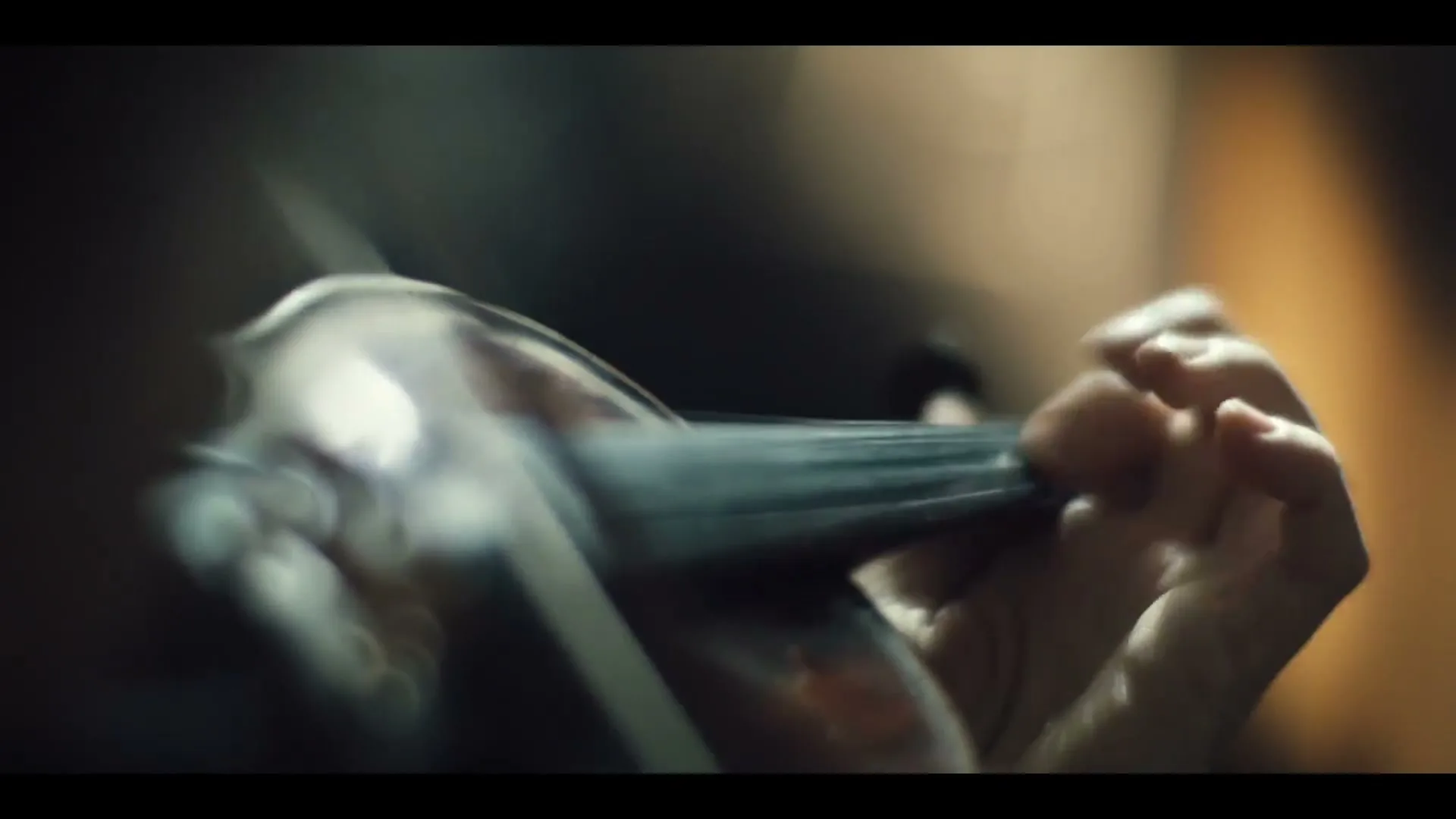 A person's hand holds a violin bow, playing the instrument in a close-up shot, with soft lighting.