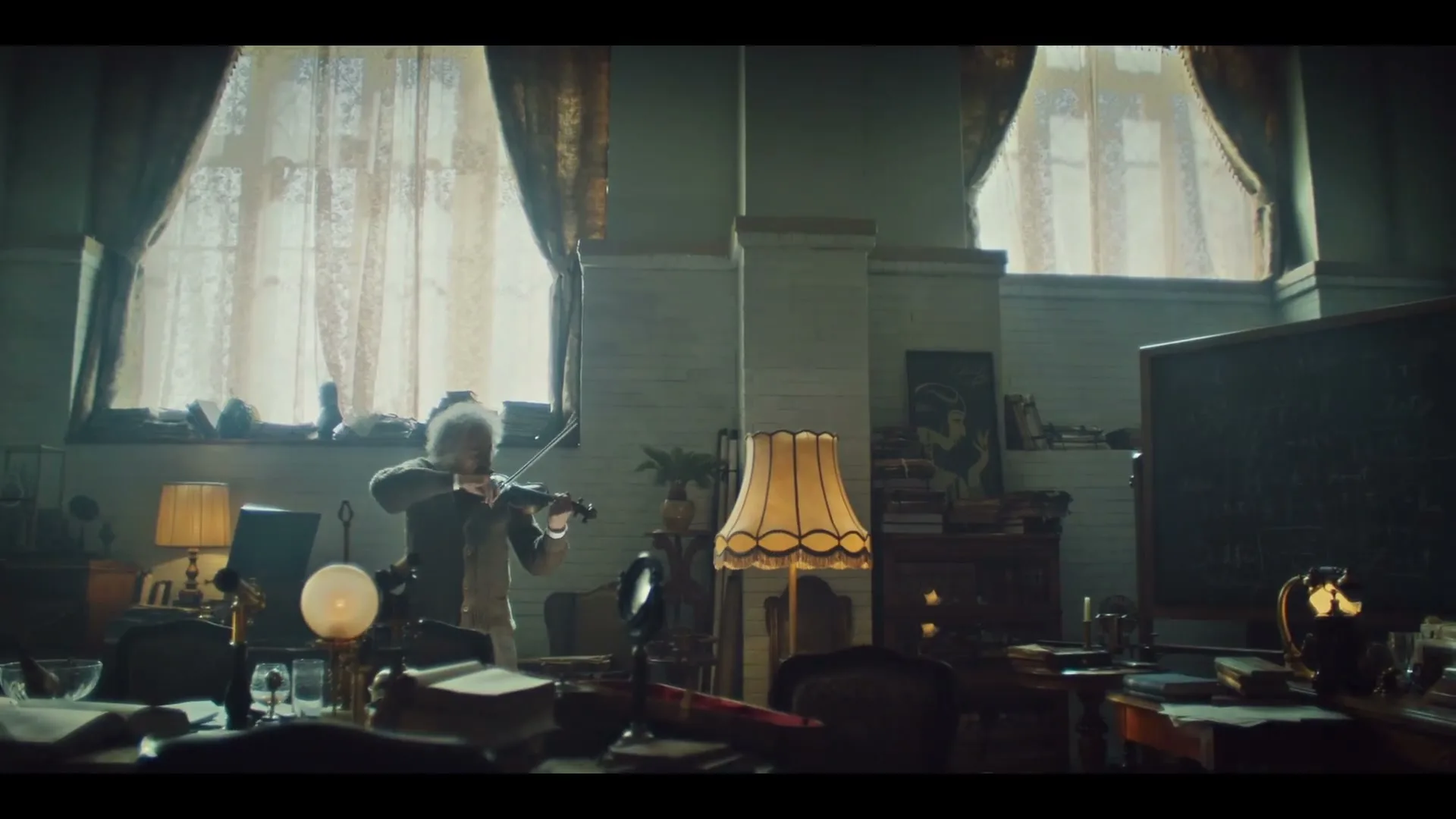 An older man with white hair, dressed in a suit, plays the violin in a cluttered office with large windows and a blackboard.