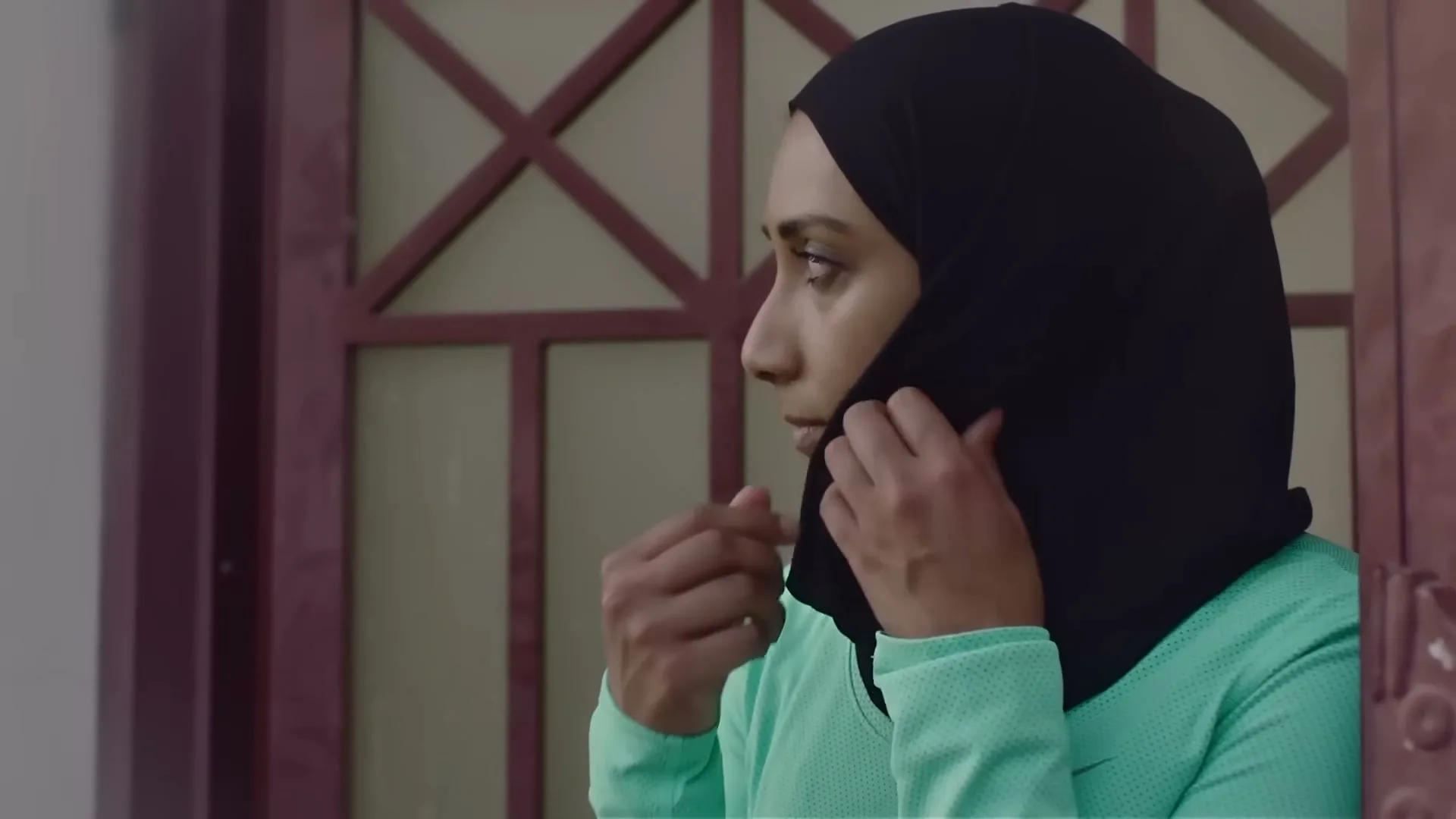 A young woman in a hijab and green athletic top peeks out from behind a gate, then walks away down a street.