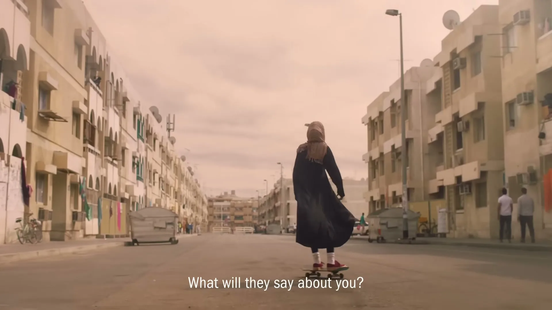 A young woman in a hijab and abaya skateboards down a street in a residential area, with buildings lining both sides.