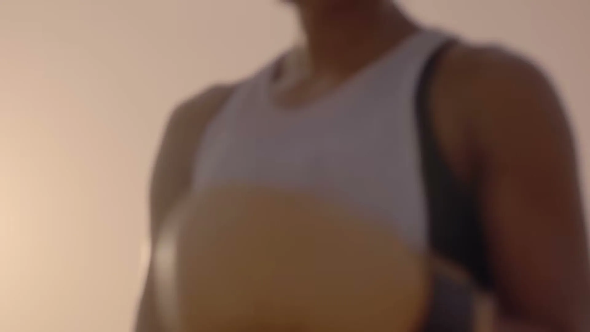 A female boxer in a white tank top and black sports bra trains in a boxing ring, with a blurred background of a young boy watching.