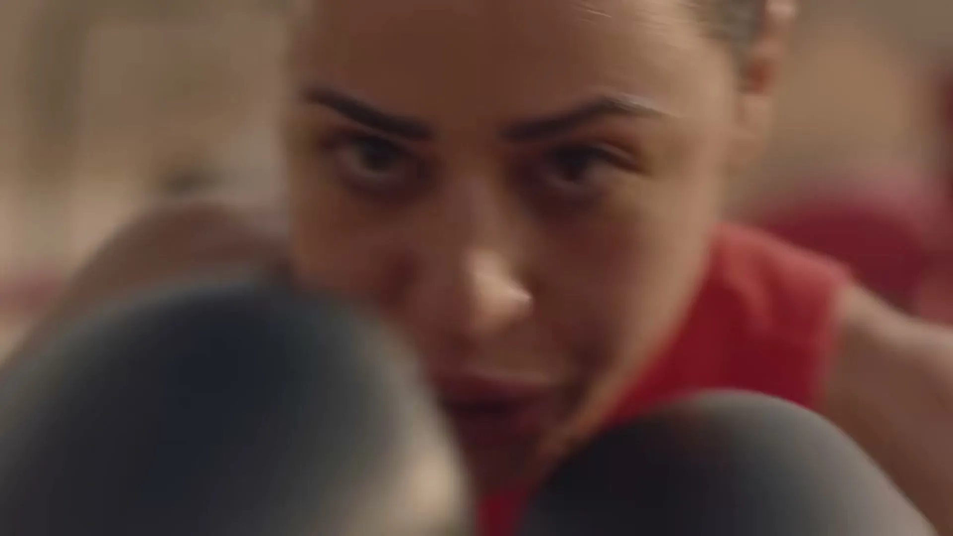 A female boxer in a red top and black gloves trains intensely, looking directly at the camera in a boxing ring.