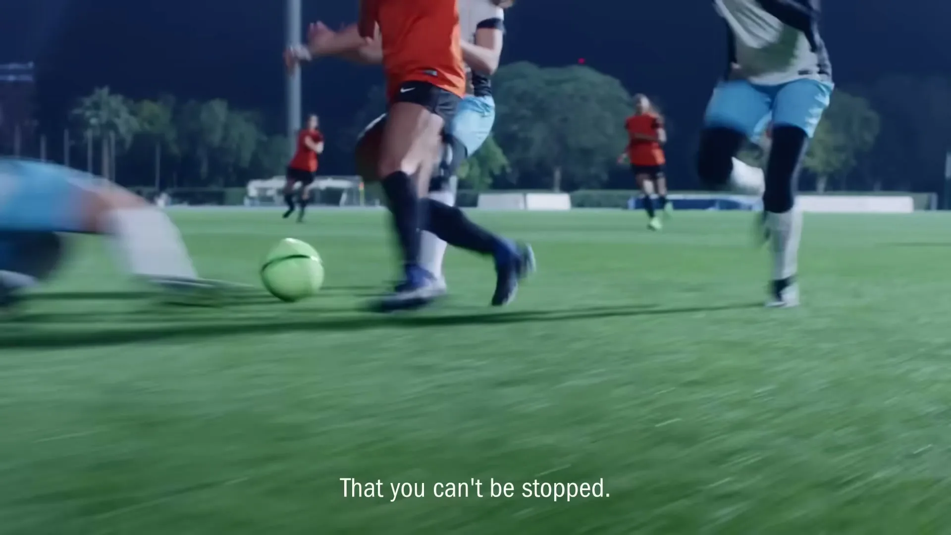 A female soccer player in an orange jersey dribbles a neon green ball down a stadium field at night, with other players running around her.