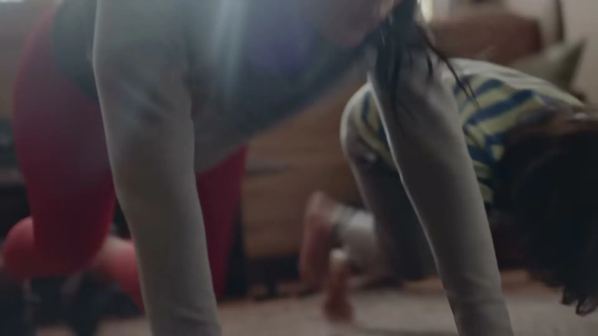 A woman and a child are doing bear crawls on a patterned rug in a sunlit room, with a smartphone lying on the floor between them.