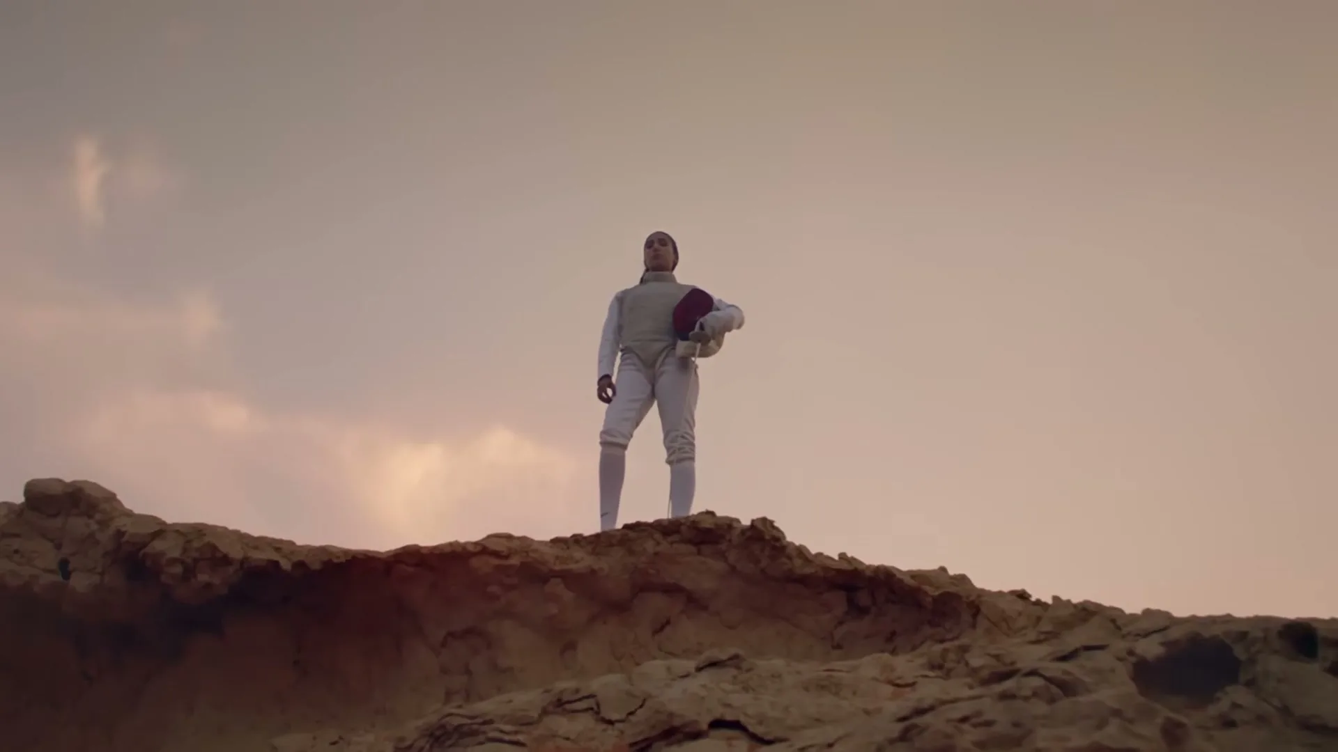 A female fencer in white uniform and hijab stands triumphantly on a rocky hill, holding her helmet, against a dramatic sky.