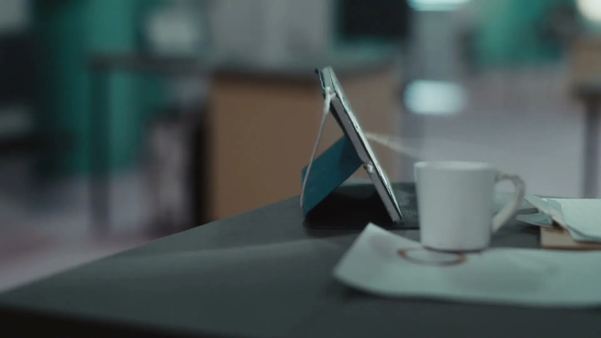 A tablet on a stand and a white coffee cup sit on a dark desk in an office, with a hand reaching in to unplug a cable.