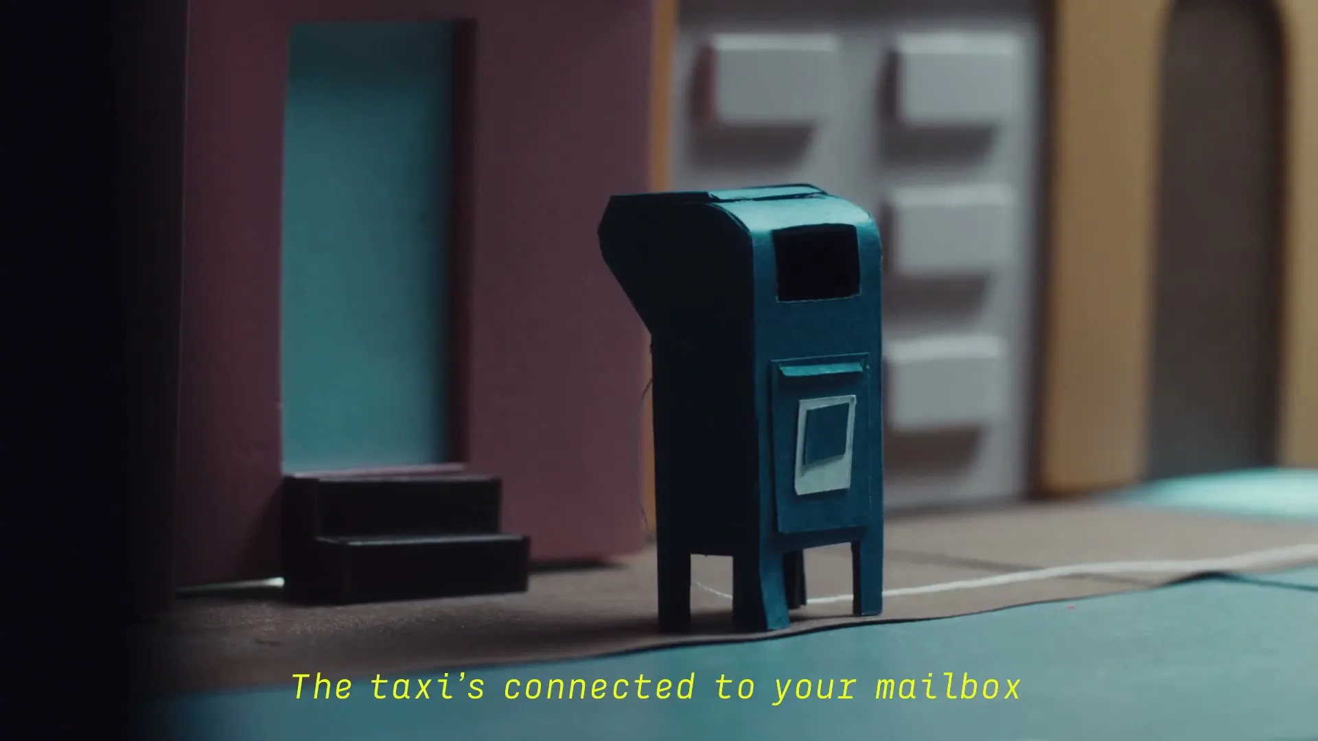 A miniature yellow taxi drives past a blue mailbox on a street made of cardboard, with a building in the background.