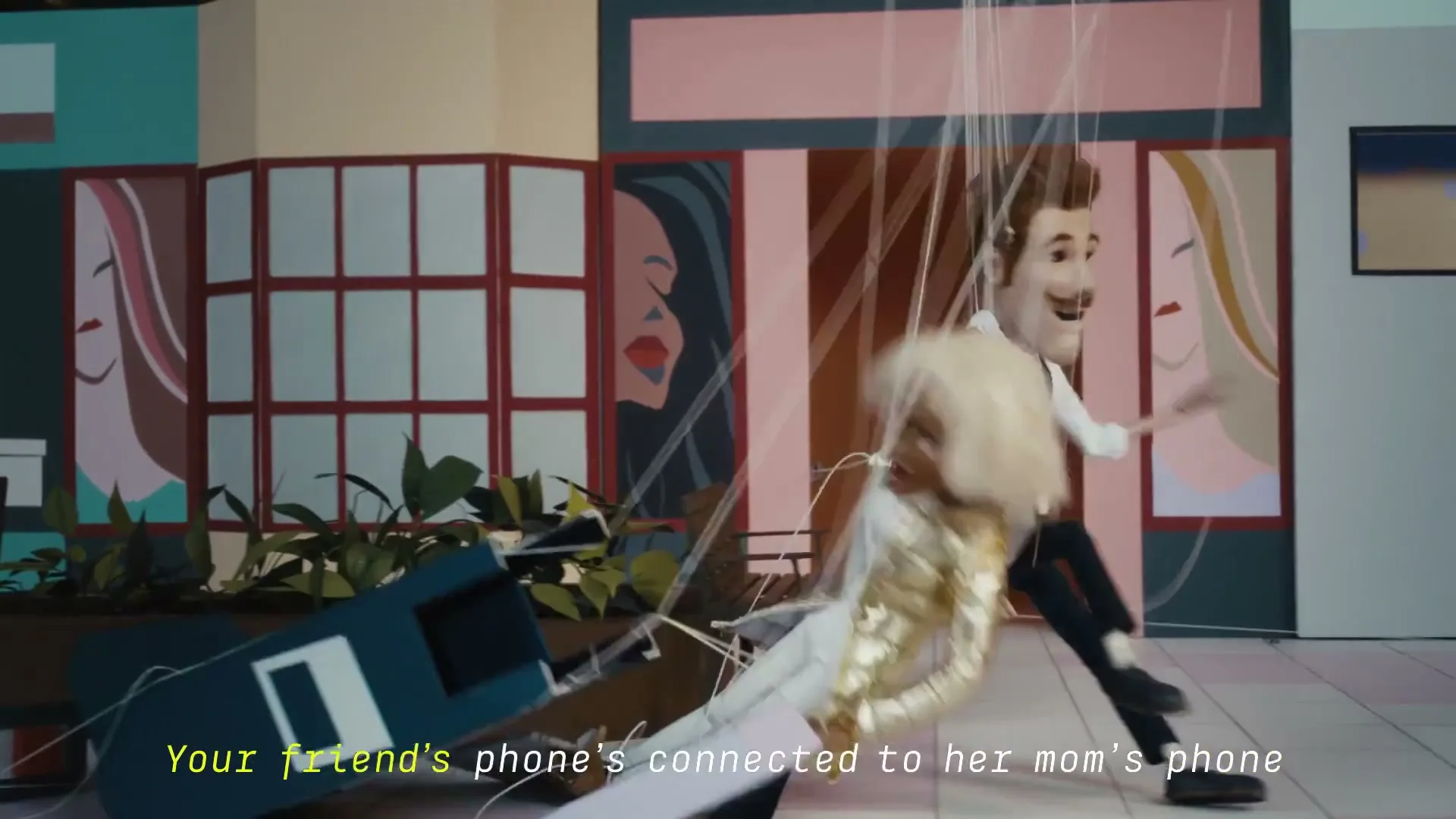 A male puppet in a white shirt and suspenders dances with a female puppet in a gold jumpsuit, both controlled by strings, in a colorful, stylized interior.