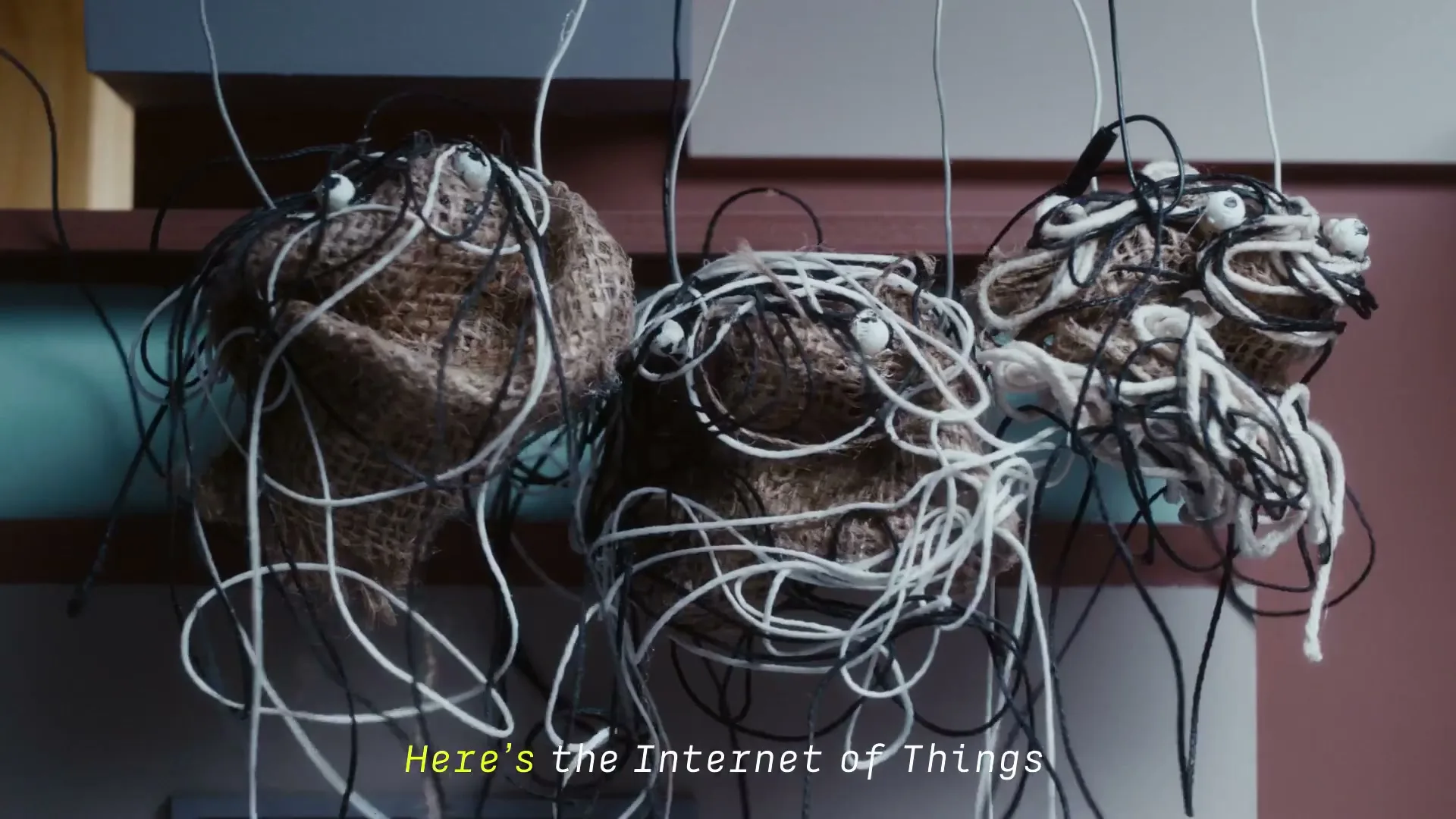 Three brown, burlap-like puppets with tangled black and white wires and googly eyes hang from above, with a subtitle at the bottom of the screen.