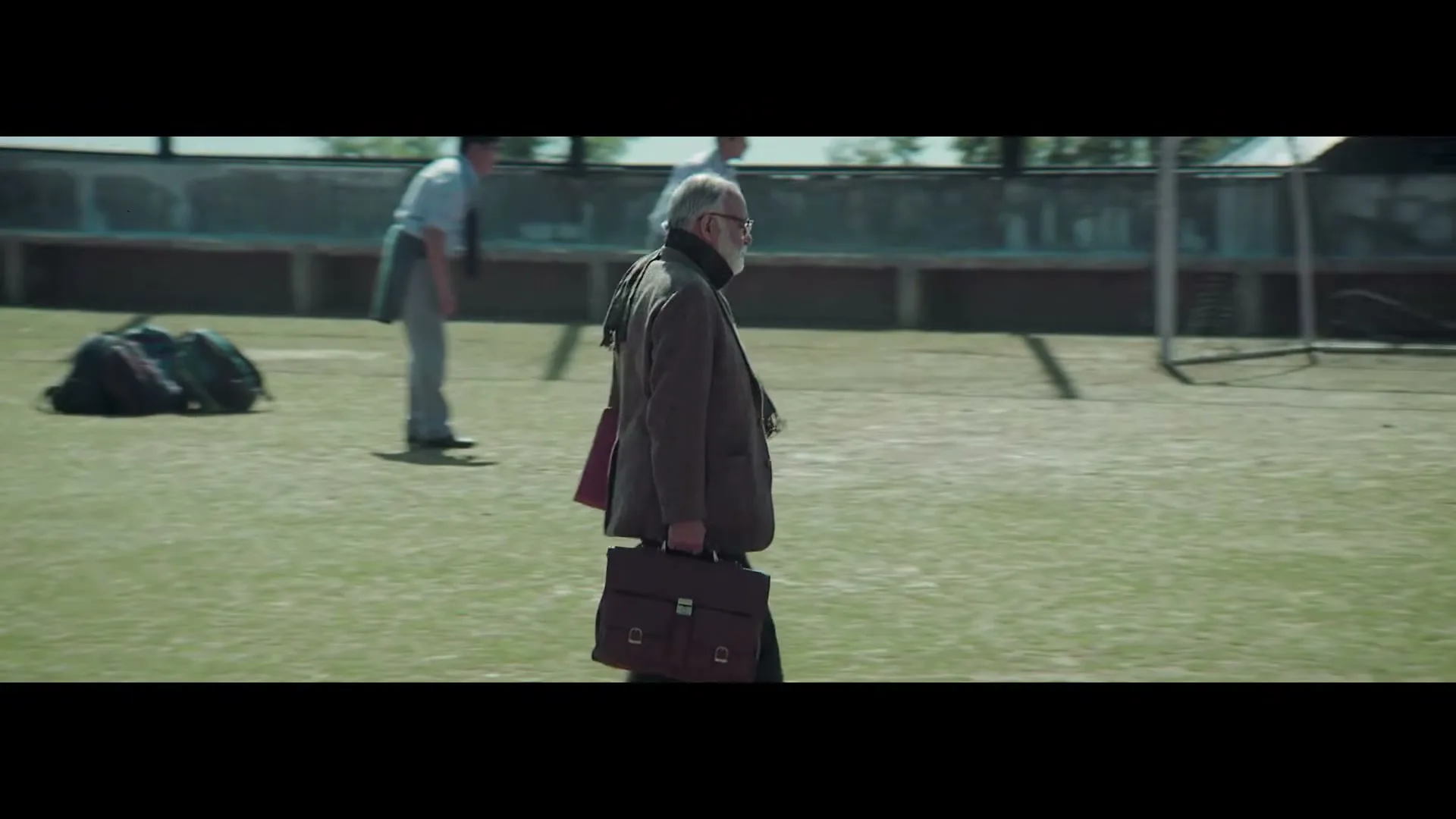 An older man with a white beard and glasses walks across a green sports field, carrying a briefcase, as younger men play soccer in the background.