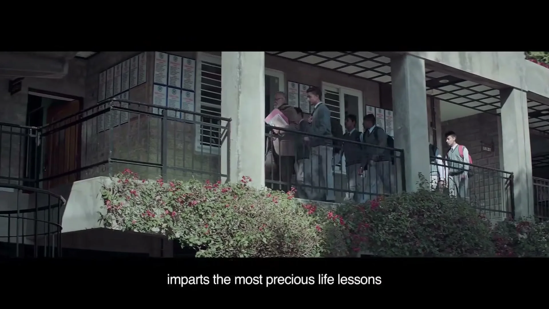 A male teacher in a brown suit walks out of a building with students in school uniforms following him, on a sunny day.