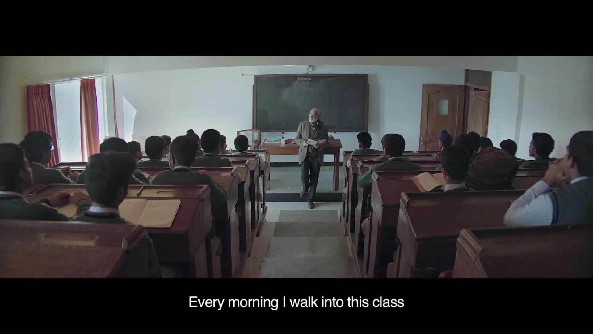 An elderly male teacher with a white beard stands in front of a classroom of young male students, holding a book, in a wide shot.