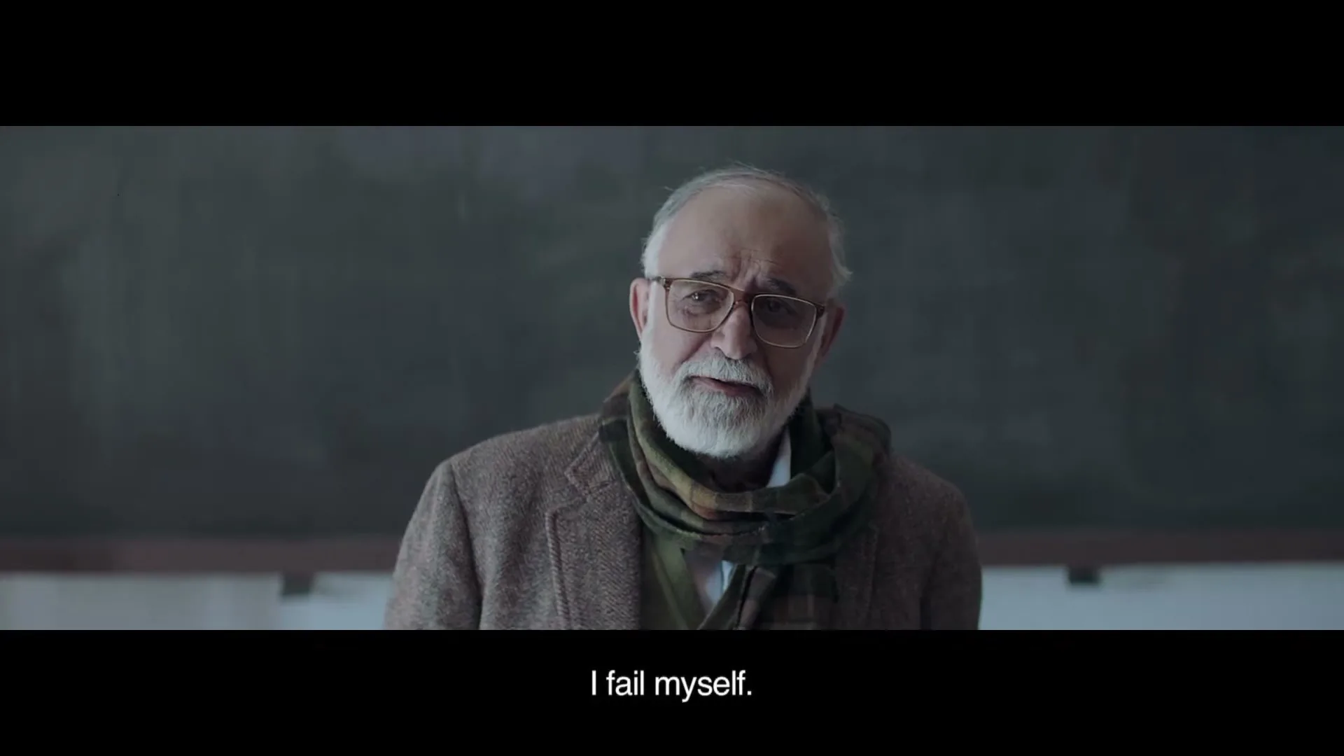 An elderly man with a white beard and glasses, wearing a brown jacket and scarf, looks down sadly in front of a blackboard.