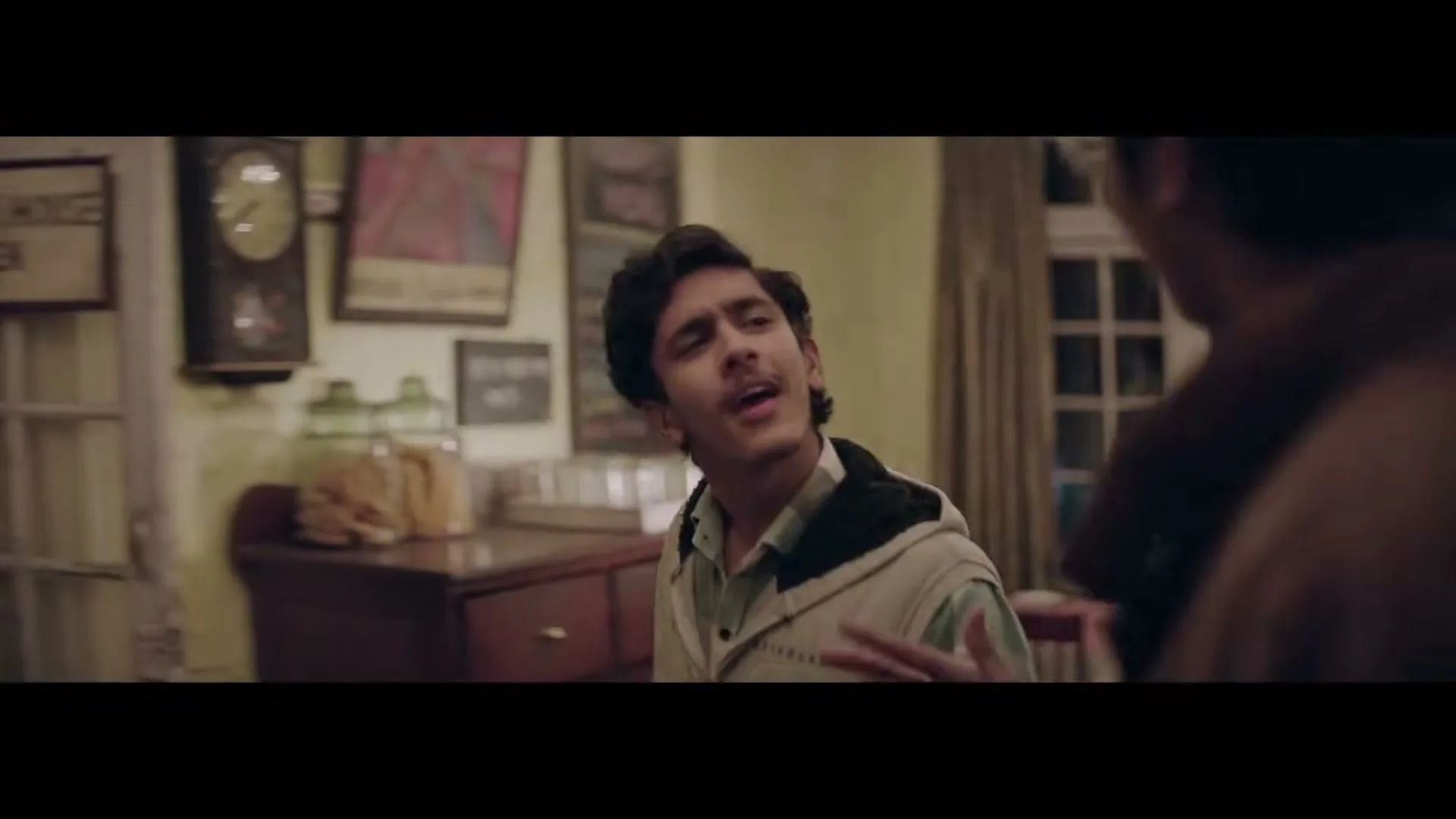 A young man in a light jacket talks animatedly to another man in a cozy, dimly lit coffee house at night.