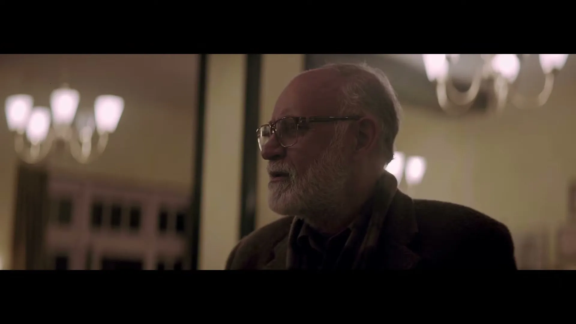 A kind older man with a white beard and glasses looks around a warmly lit interior, then looks down, in a medium shot.