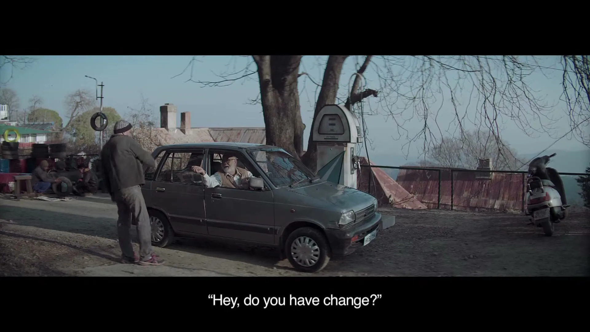 A man in a grey car asks a gas station attendant for change, who gestures that he doesn't have any, at an outdoor gas station.