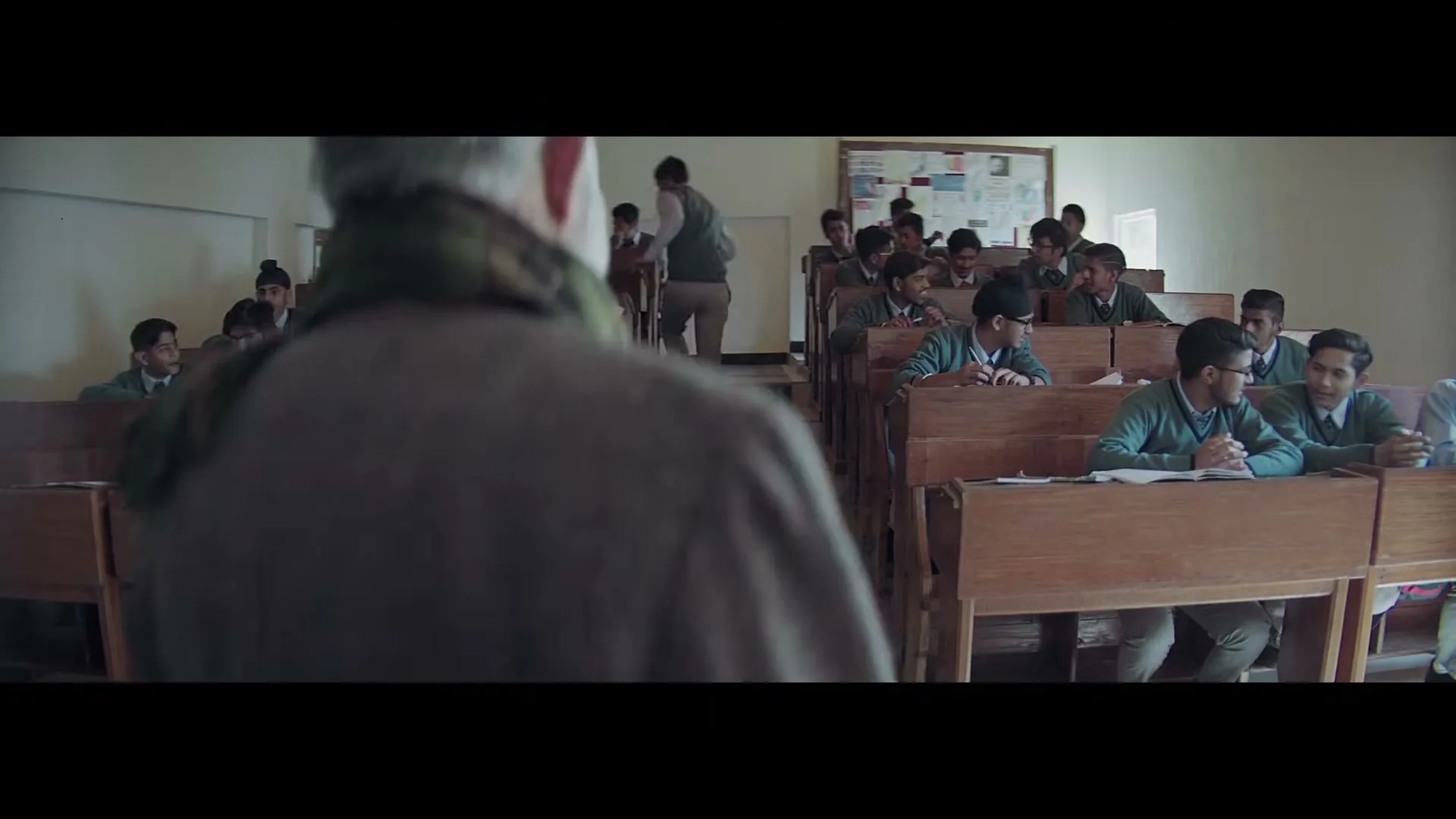A male teacher with grey hair and a scarf walks into a classroom full of male students in uniforms, who are seated at wooden desks.