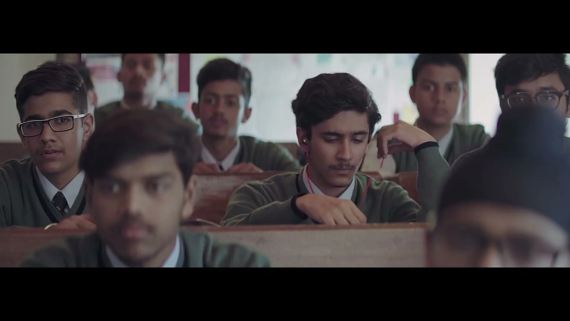 A male student in a green sweater and white shirt with a tie, wearing earphones, looks down in a classroom full of other students.