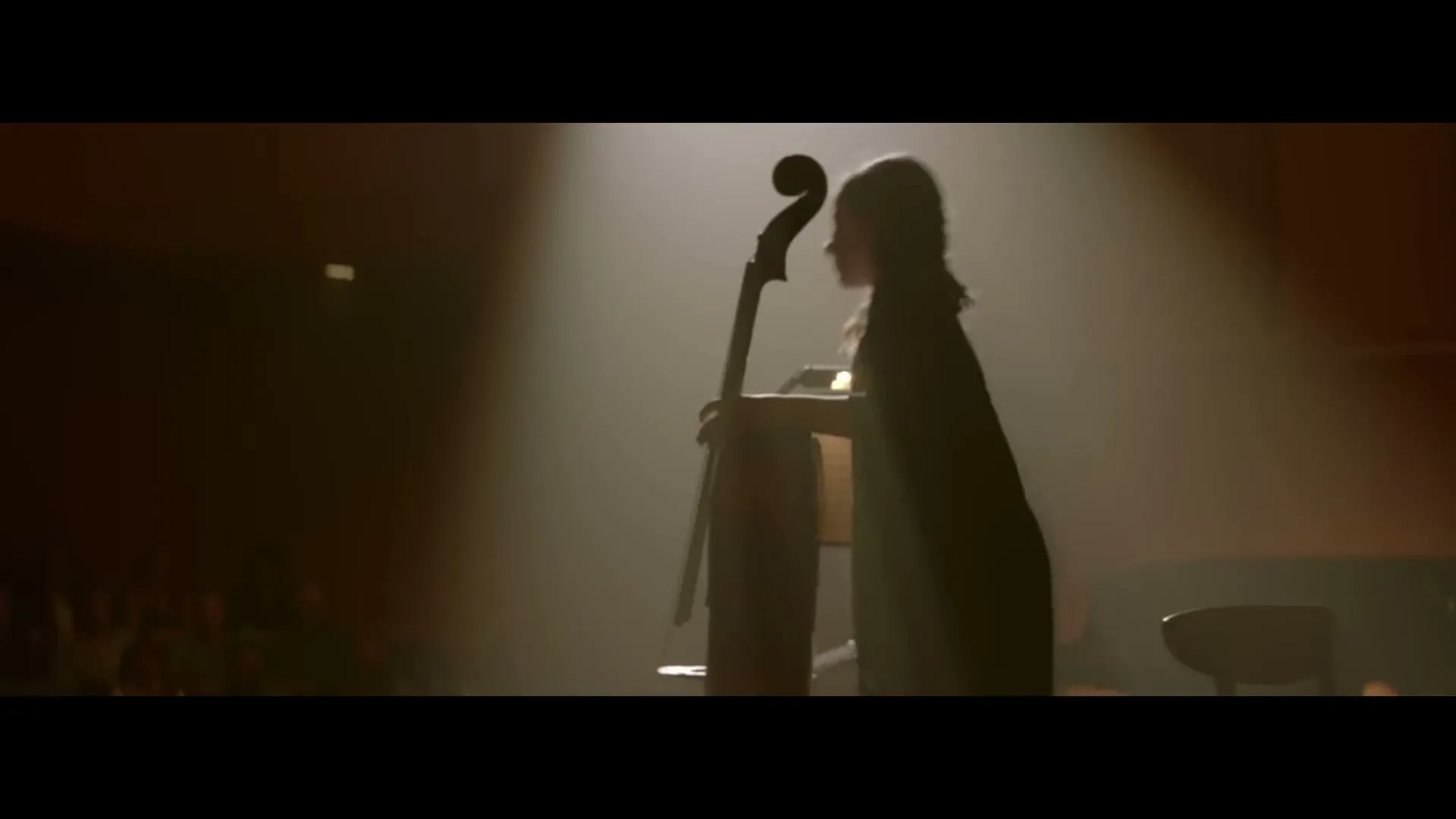 A young woman in a dark dress stands on a dimly lit stage, holding an upright bass, preparing to play for an unseen audience.