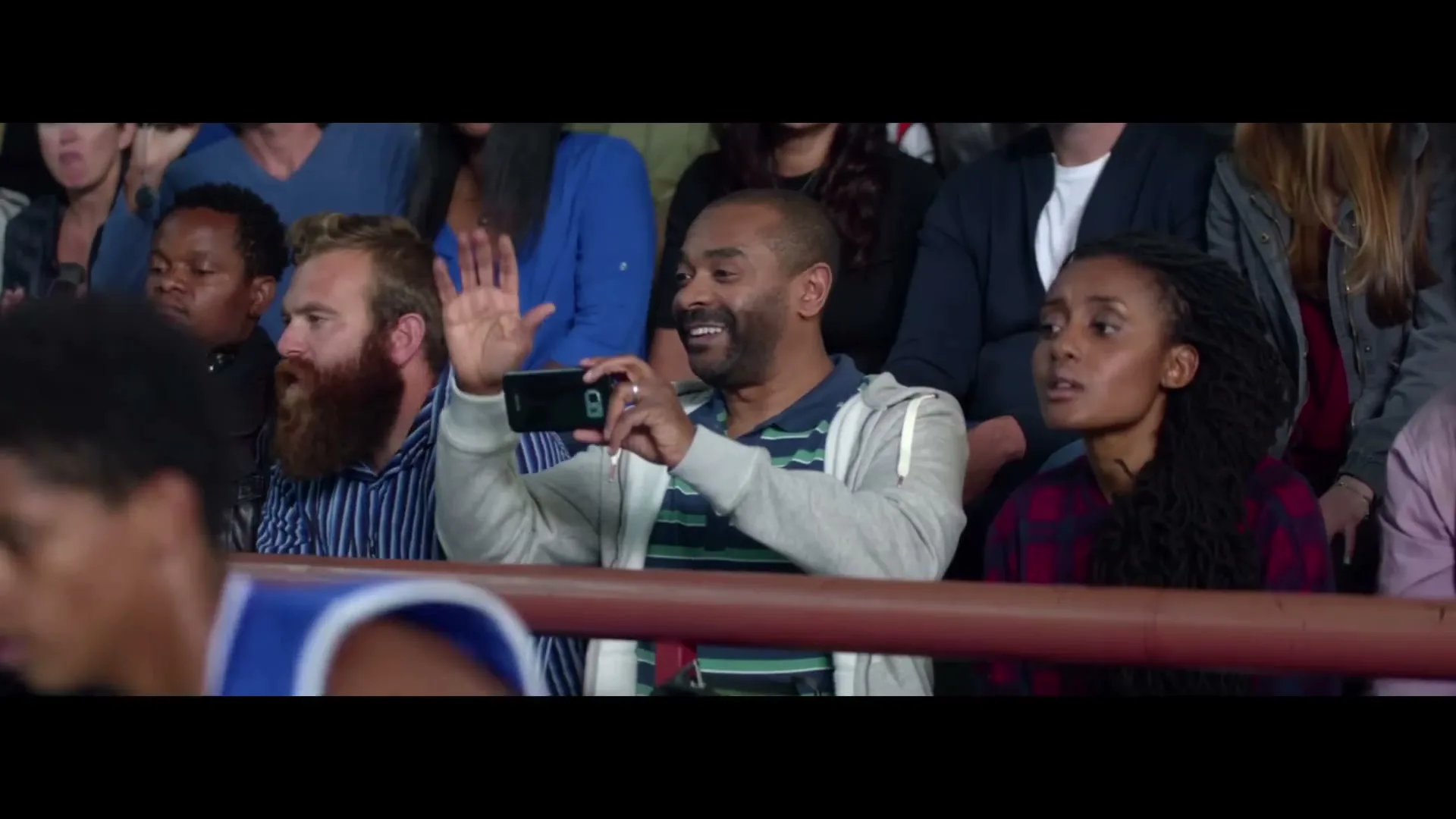 A diverse group of adults sits in stadium seats, watching an event. One man in the center holds up a smartphone to record, while another man with a beard waves.