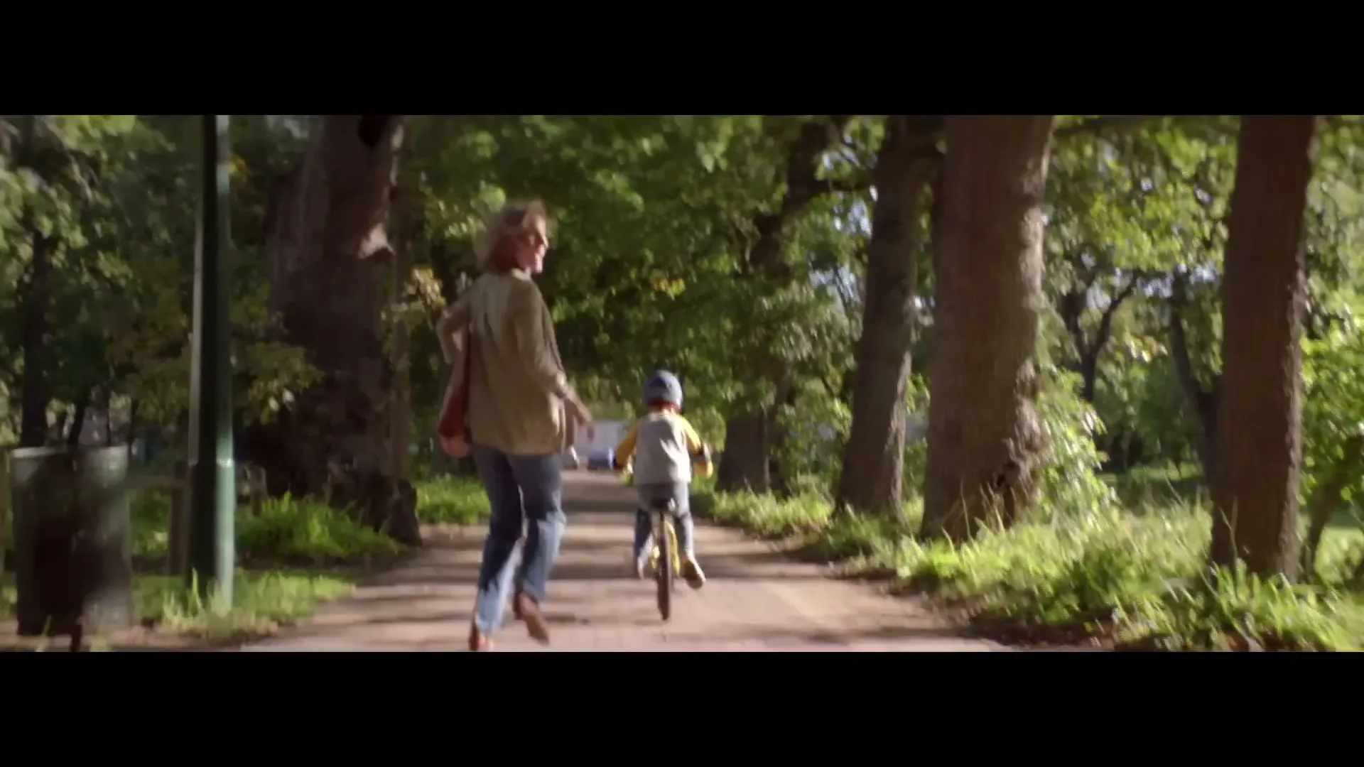 A woman in a beige jacket and jeans runs alongside a young boy on a bicycle, both moving away from the camera on a tree-lined path in a park.