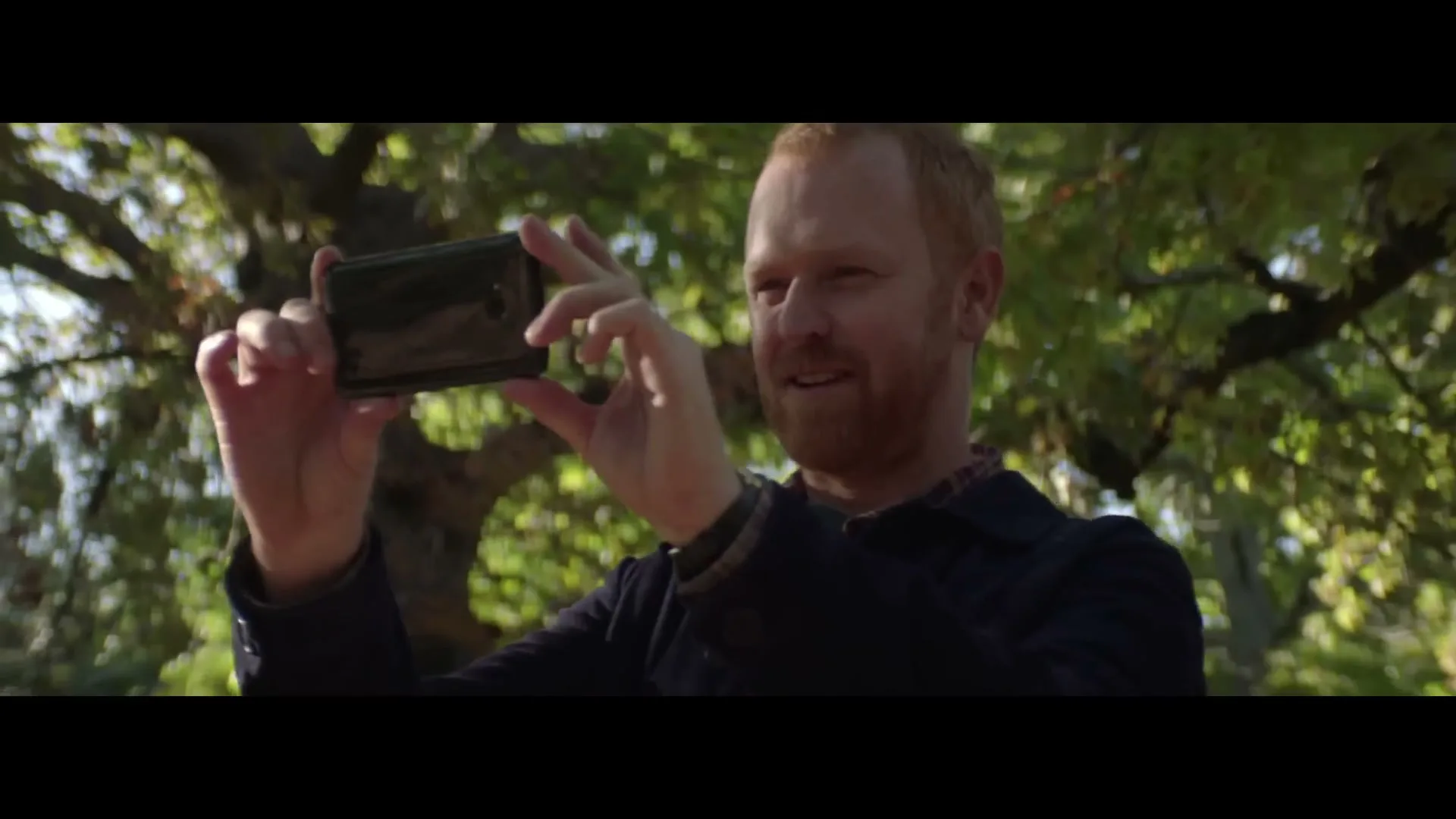 A red-haired man with a beard smiles while holding a smartphone horizontally, seemingly recording or taking a photo outdoors with trees in the background.